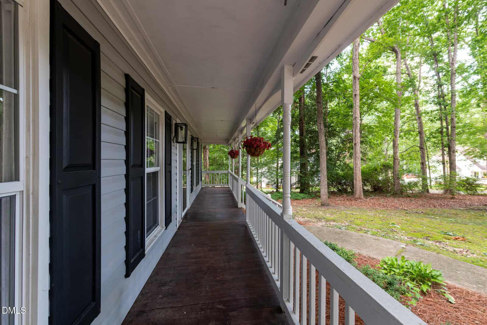 A wooden porch with white railings and hanging flower baskets overlooks a yard with trees and greenery on a sunny day.