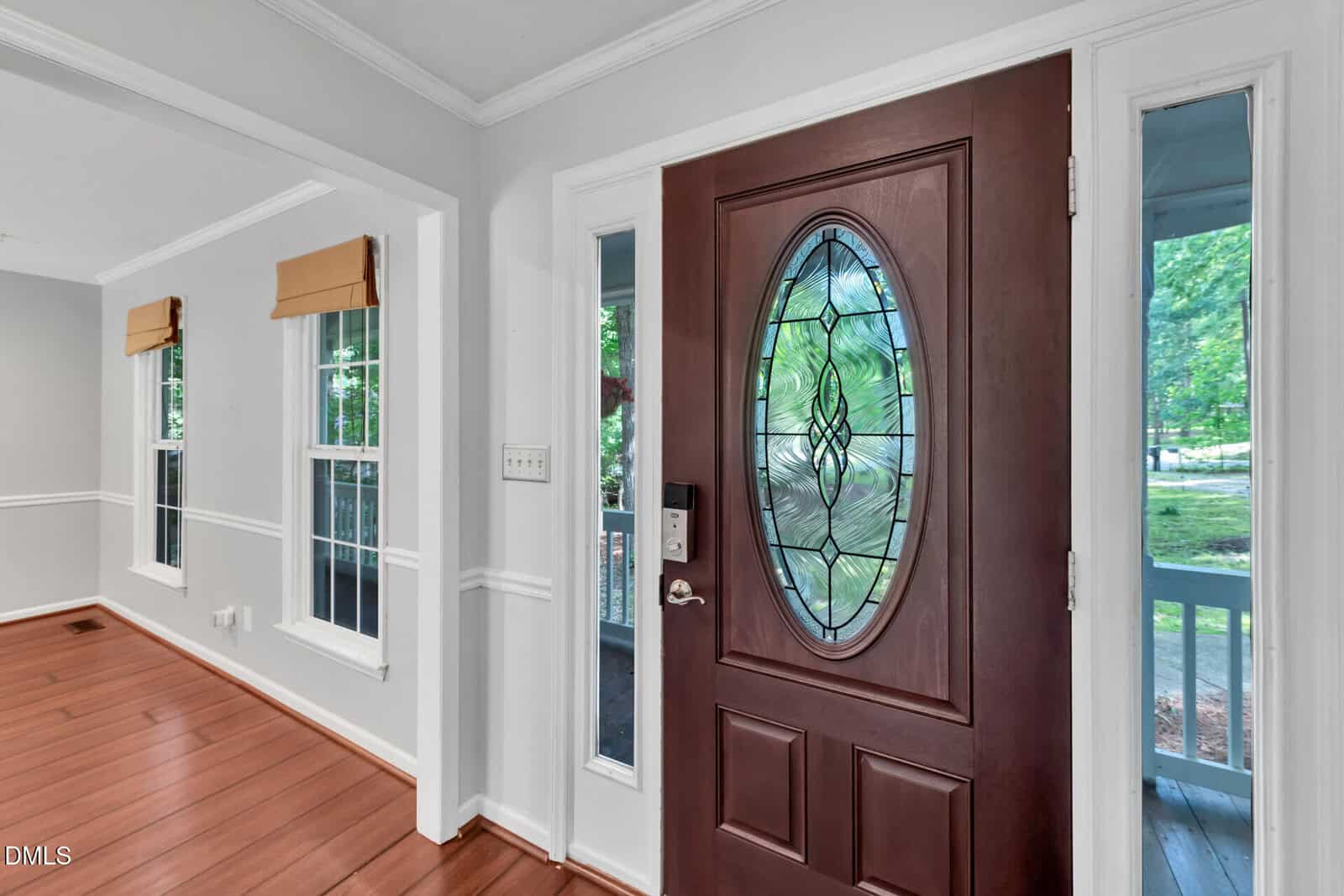 A front door with an oval stained-glass window is open, revealing a view of a green yard. Adjacent windows let in natural light, and the interior has wood floors and white trim.