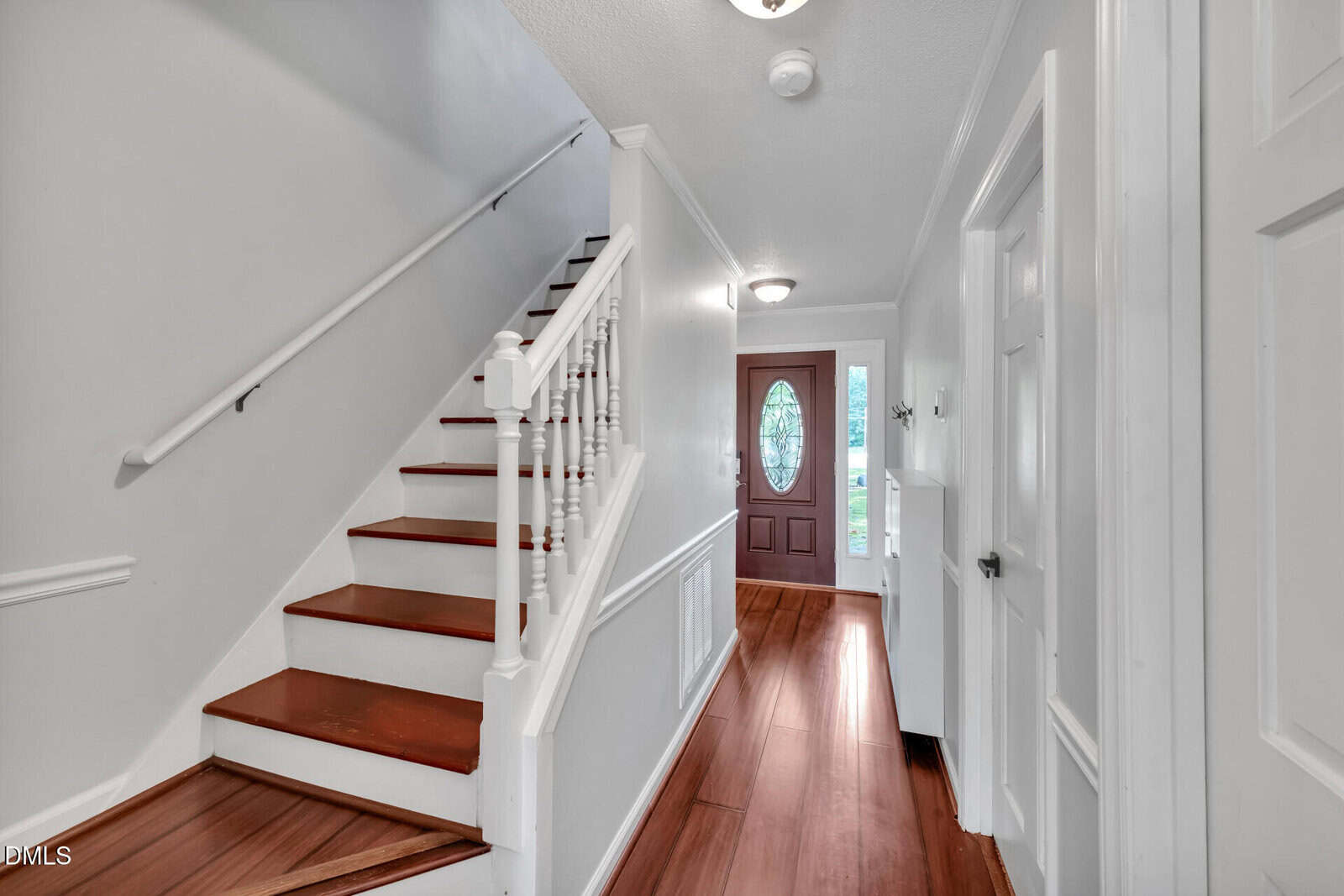 A hallway with wooden floors, white walls, and a staircase with white railings. There is a wooden front door with a decorative window at the far end, and several doors along the hallway.