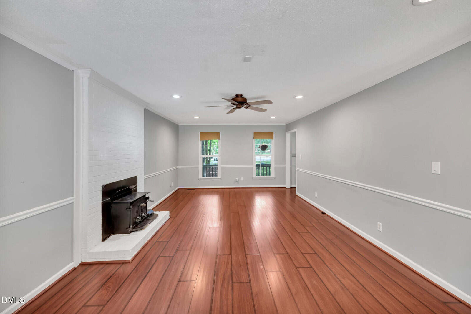 Spacious living room with wood flooring, gray walls, white trim, a ceiling fan, and a white painted brick fireplace. Two large windows with blinds face the backyard, letting in natural light.