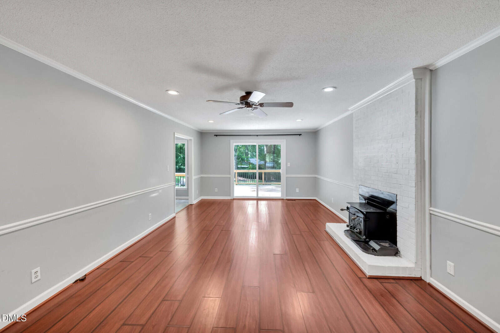 Spacious living room with wood flooring, gray walls, ceiling fan, recessed lighting, large window, sliding glass door, and a white painted brick fireplace with a black wood stove.