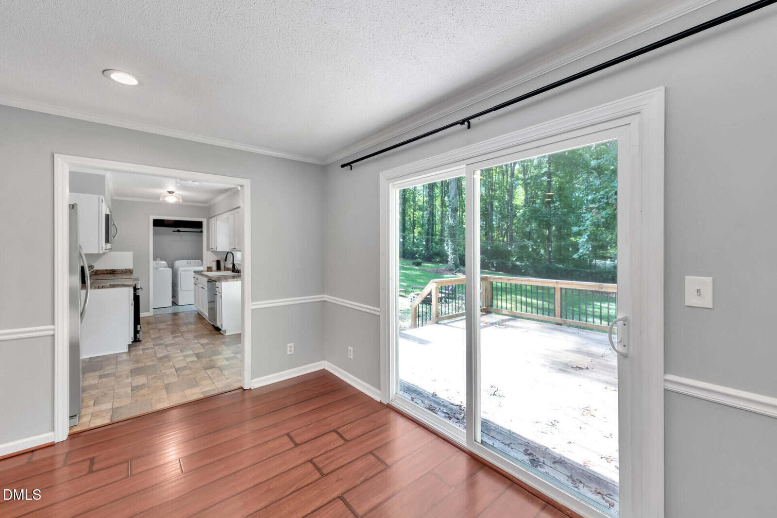 A bright room with wood floors, gray walls, and white trim. Large sliding glass doors open to a deck with a view of a green backyard. Kitchen is visible through an open doorway.