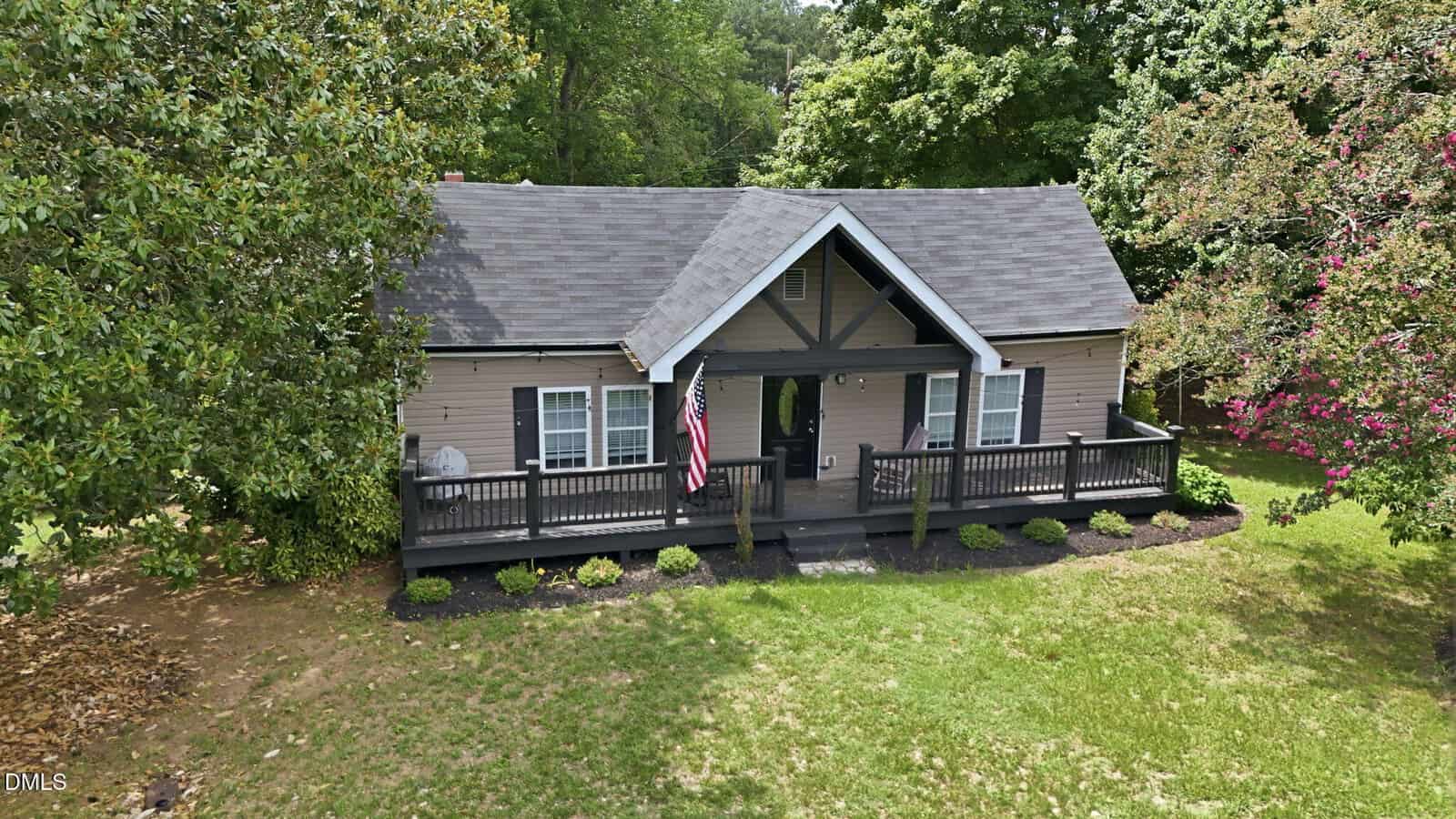 A small brown house with a front porch, black trim, gabled roof, American flag, and neatly trimmed shrubs, surrounded by green trees and a grassy lawn.
