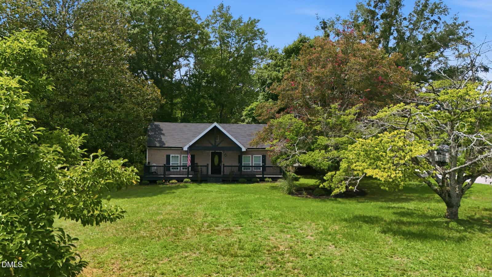 A small tan house with a dark roof sits in the center, surrounded by lush green grass and trees on a sunny day with a clear blue sky.