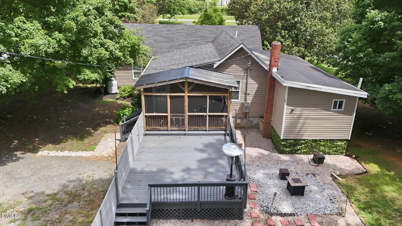 Aerial view of a house with a large backyard deck, screened porch, fire pit area, and surrounding trees and greenery.