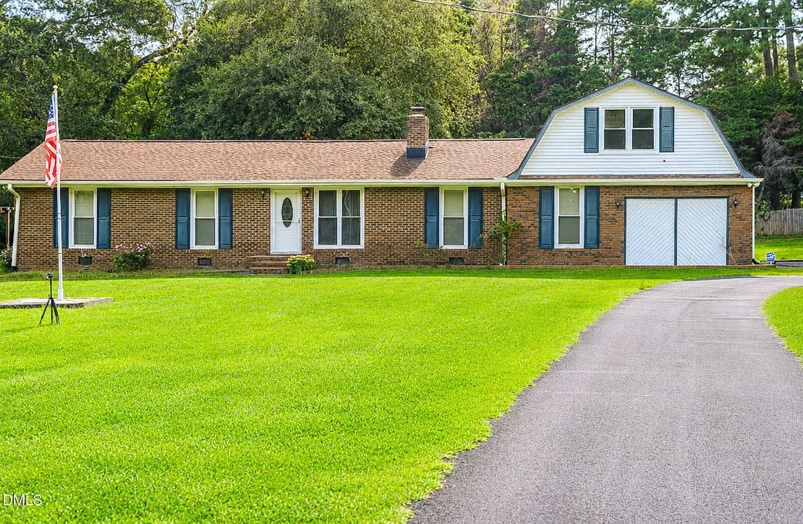 Single-story brick house with a two-story garage, blue shutters, and a well-kept lawn. An American flag stands by a curved driveway, surrounded by trees and greenery.
