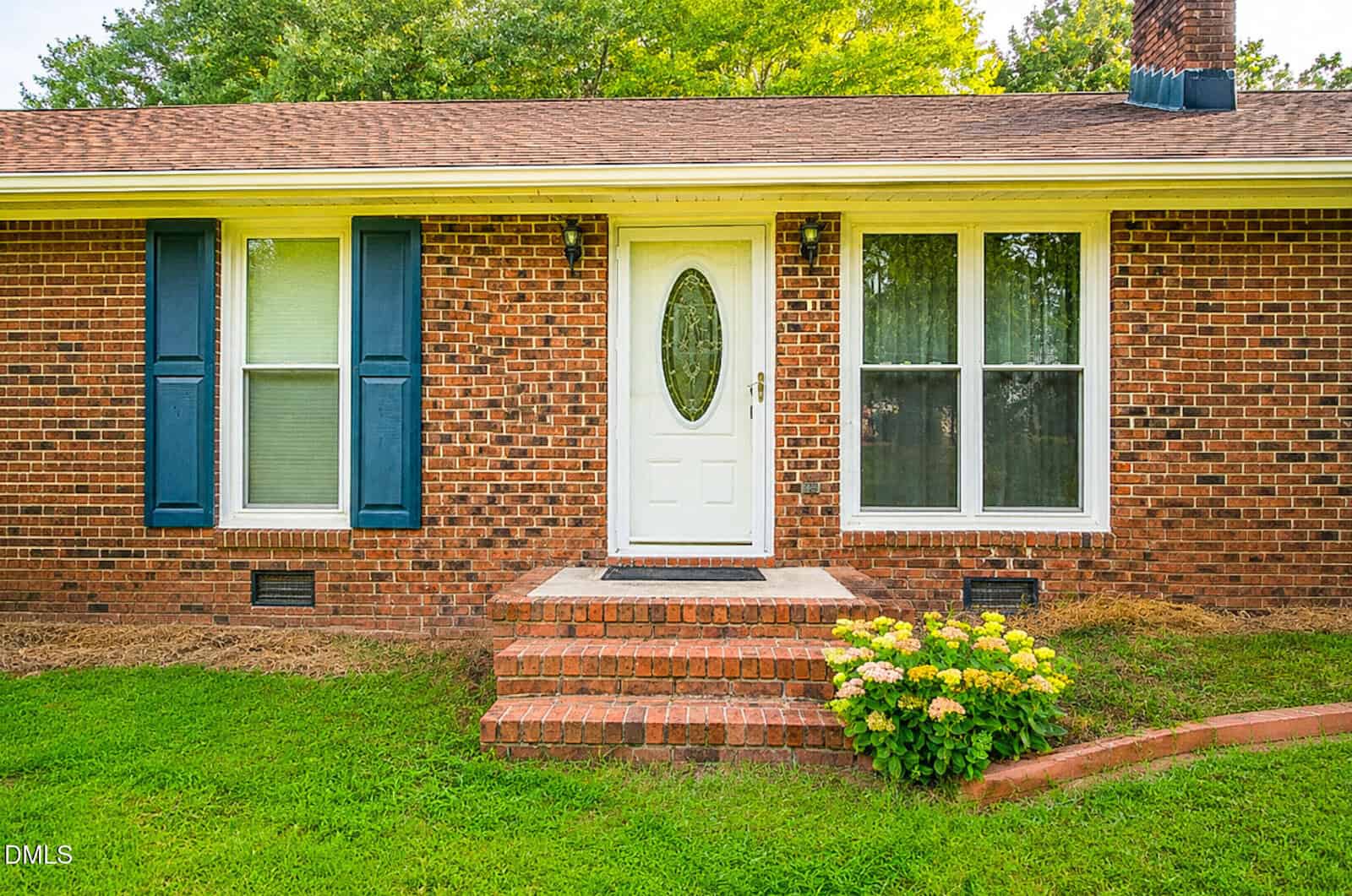 Brick house exterior with white front door, two windows with blue shutters, brick steps, green grass, small flower bush, and trees in the background.