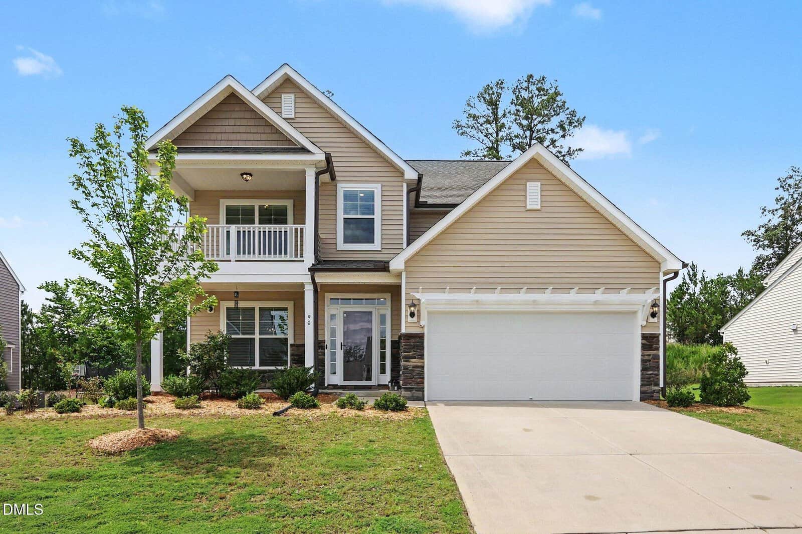 Charming two-story beige house at 90 Bourne Drive, Franklinton, featuring a welcoming front porch, balcony, white trim, and a spacious two-car garage. A small tree and well-maintained shrubs adorn the front yard, with a driveway that leads directly up to the garage.
