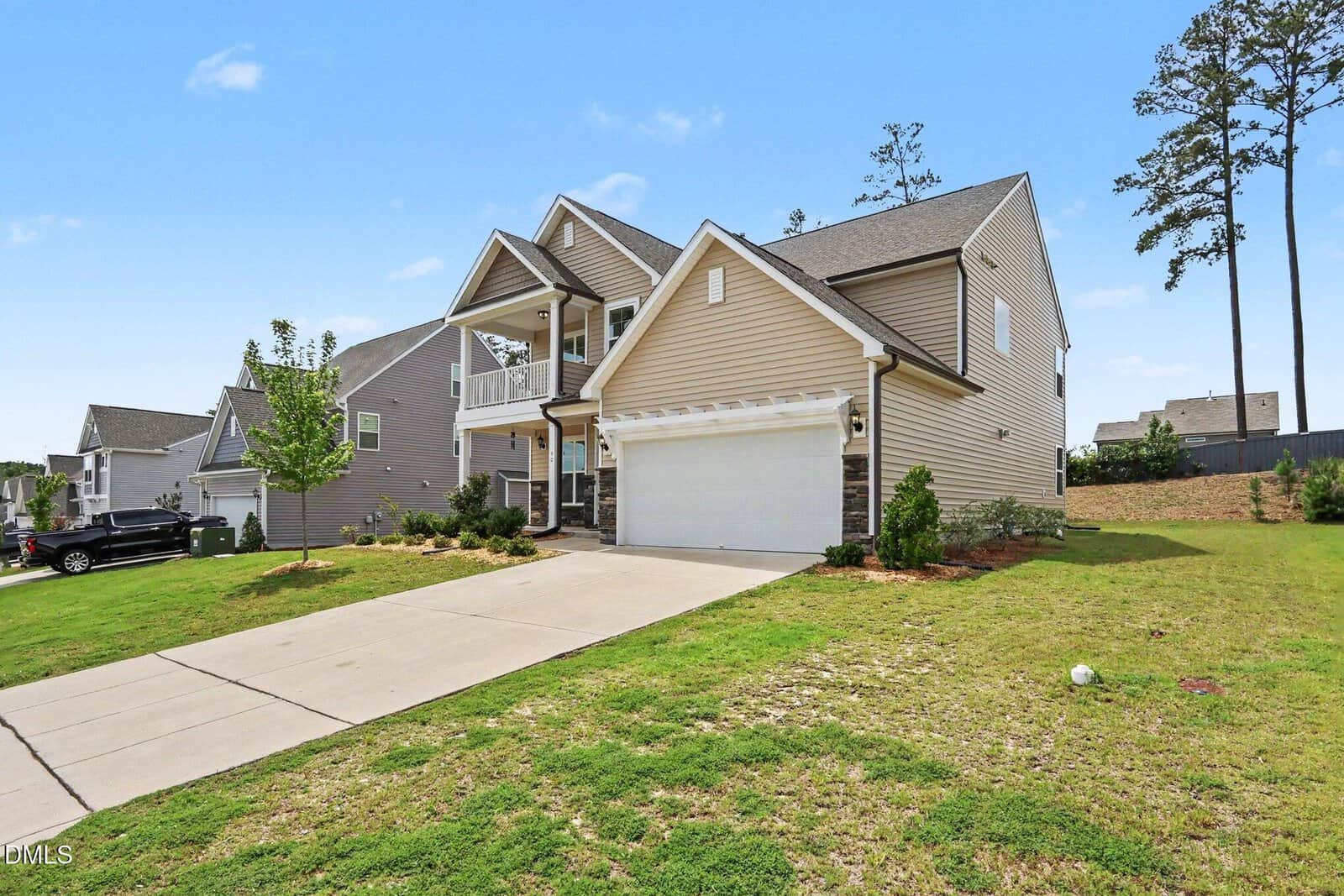 Two-story suburban house with beige siding, white trim, a double garage, and a front porch. The house sits on a green lawn with a concrete driveway and landscaping. The sky is clear and blue.