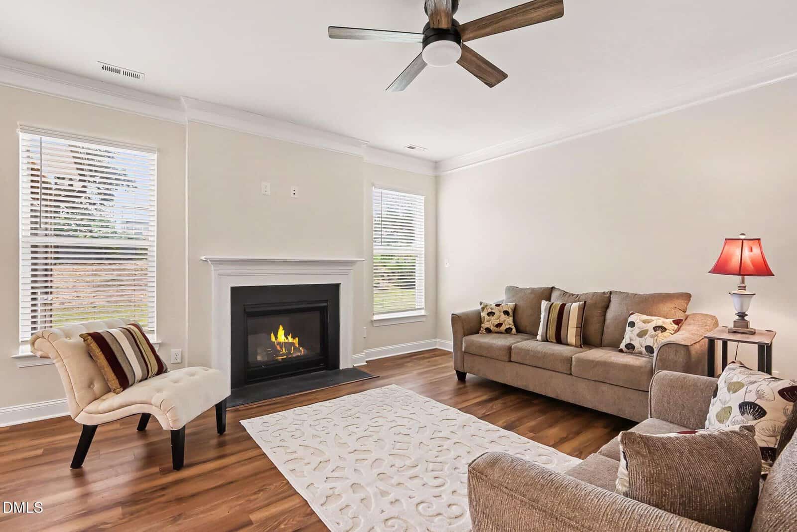 Bright living room with a ceiling fan, fireplace, beige sofa, accent chair, coffee table, and a white rug on hardwood floors. Two windows bring in natural light; a lit lamp is on an end table.