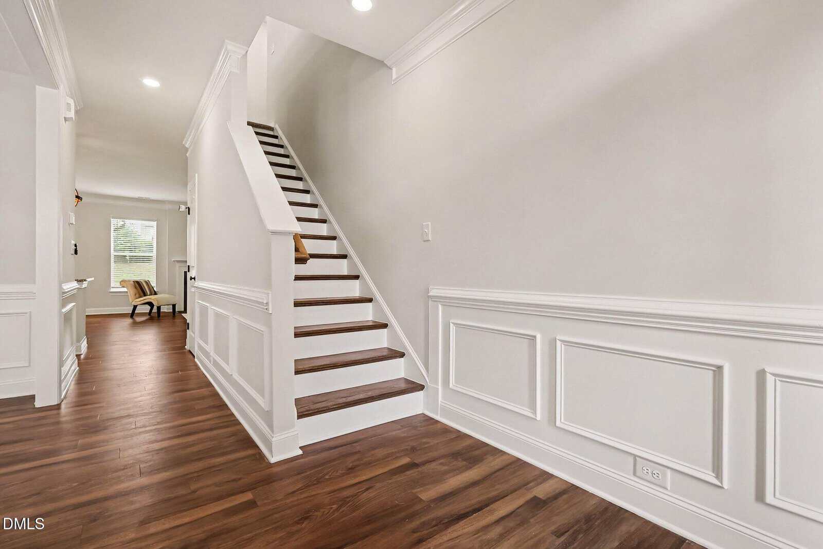A modern hallway with white paneled walls and wood flooring features a staircase with white risers and wooden steps, leading to an upper floor. Natural light enters from a window in the background.
