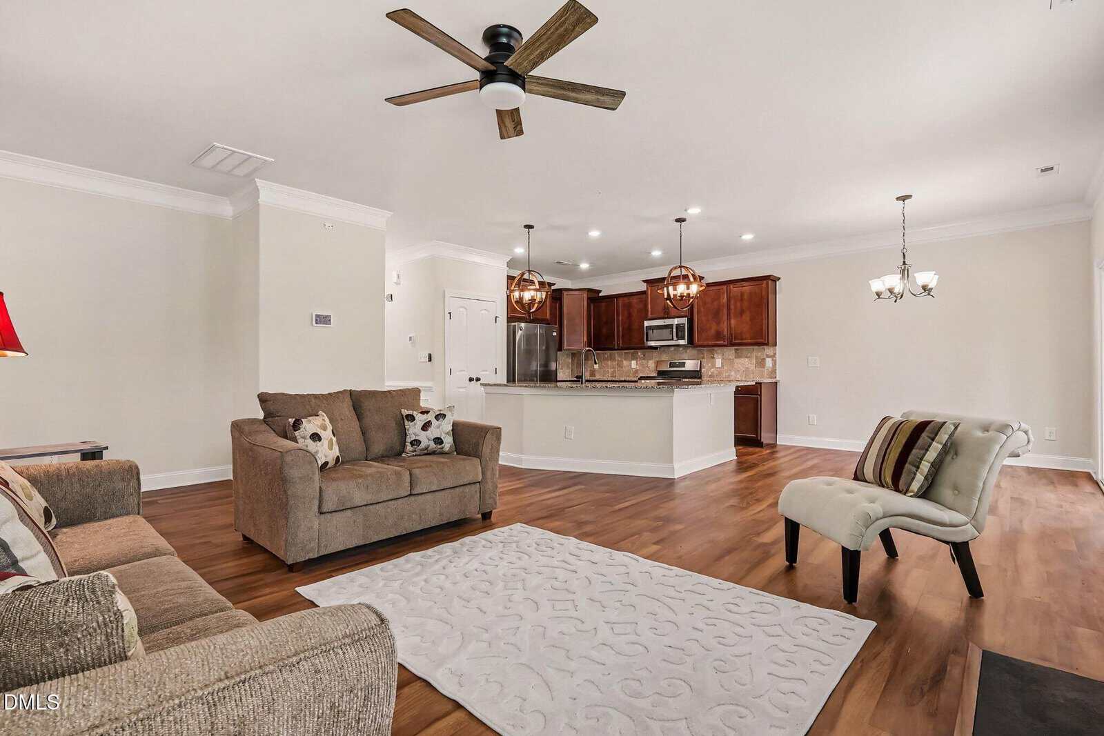 Open living room and kitchen with wood floors, brown sofas, patterned rug, white accent chair, ceiling fan, and pendant lights over a granite island and dining area with a chandelier.