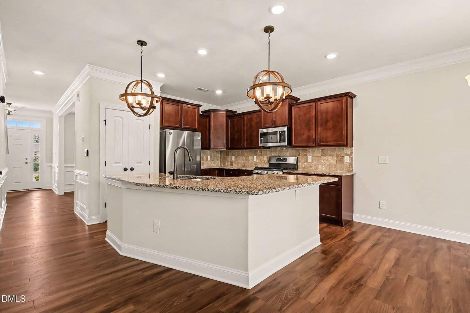 Modern kitchen with dark wood cabinets, granite countertops, stainless steel appliances, a large island with sink, pendant lights, tile backsplash, and hardwood floors. Walls are light-colored, and there’s crown molding throughout.