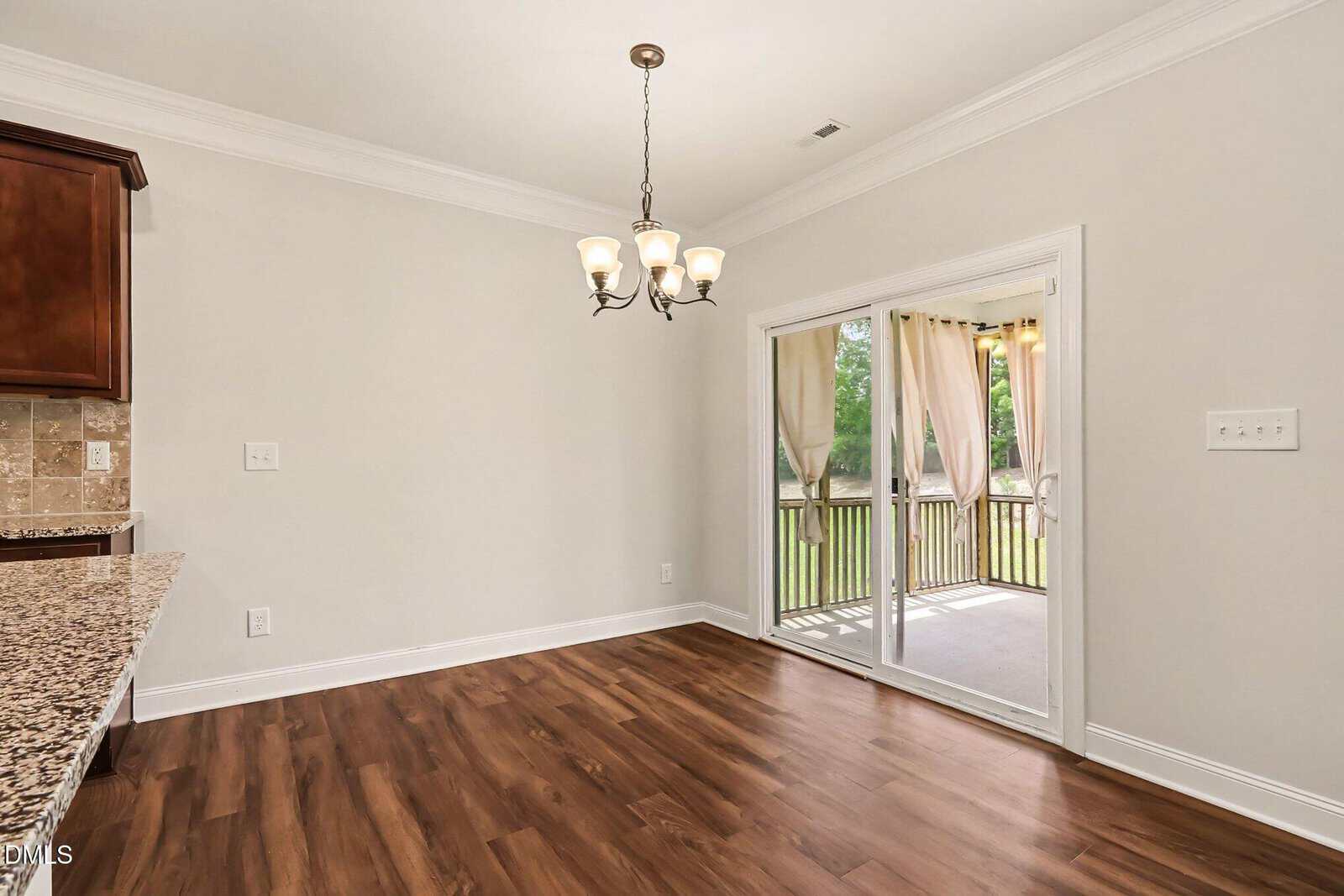 Bright dining area with wood flooring, a chandelier, and sliding glass doors leading to a covered patio with outdoor curtains and a view of greenery. Kitchen counter is partially visible on the left.