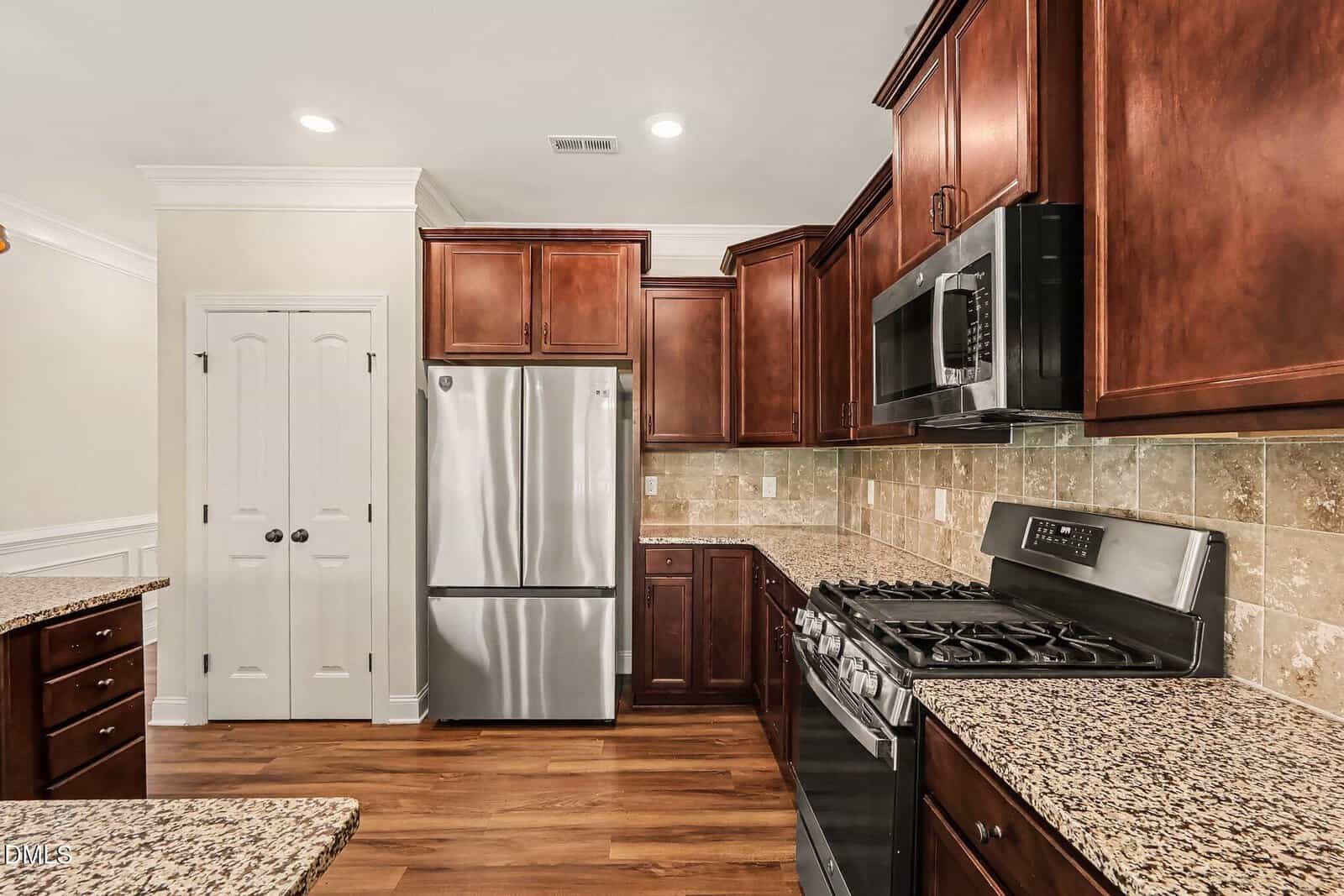 Modern kitchen with stainless steel appliances, dark wood cabinets, granite countertops, tile backsplash, and a double pantry door. Hardwood floors and recessed lighting complete the space.
