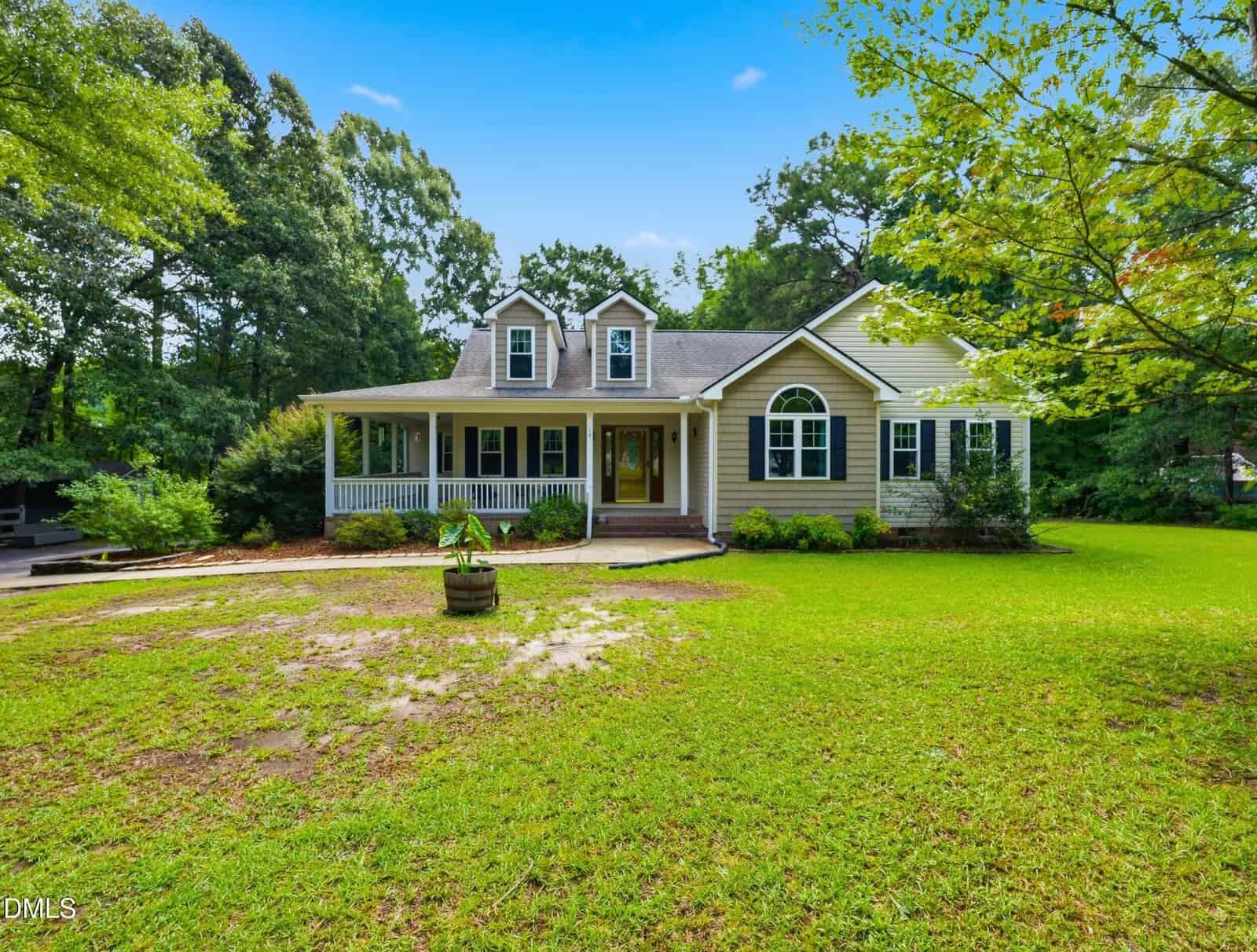 A tan house at 15 Sylvan Way in Zebulon features a large front porch, two dormer windows, and a well-kept yard, surrounded by trees and green grass under a blue sky.