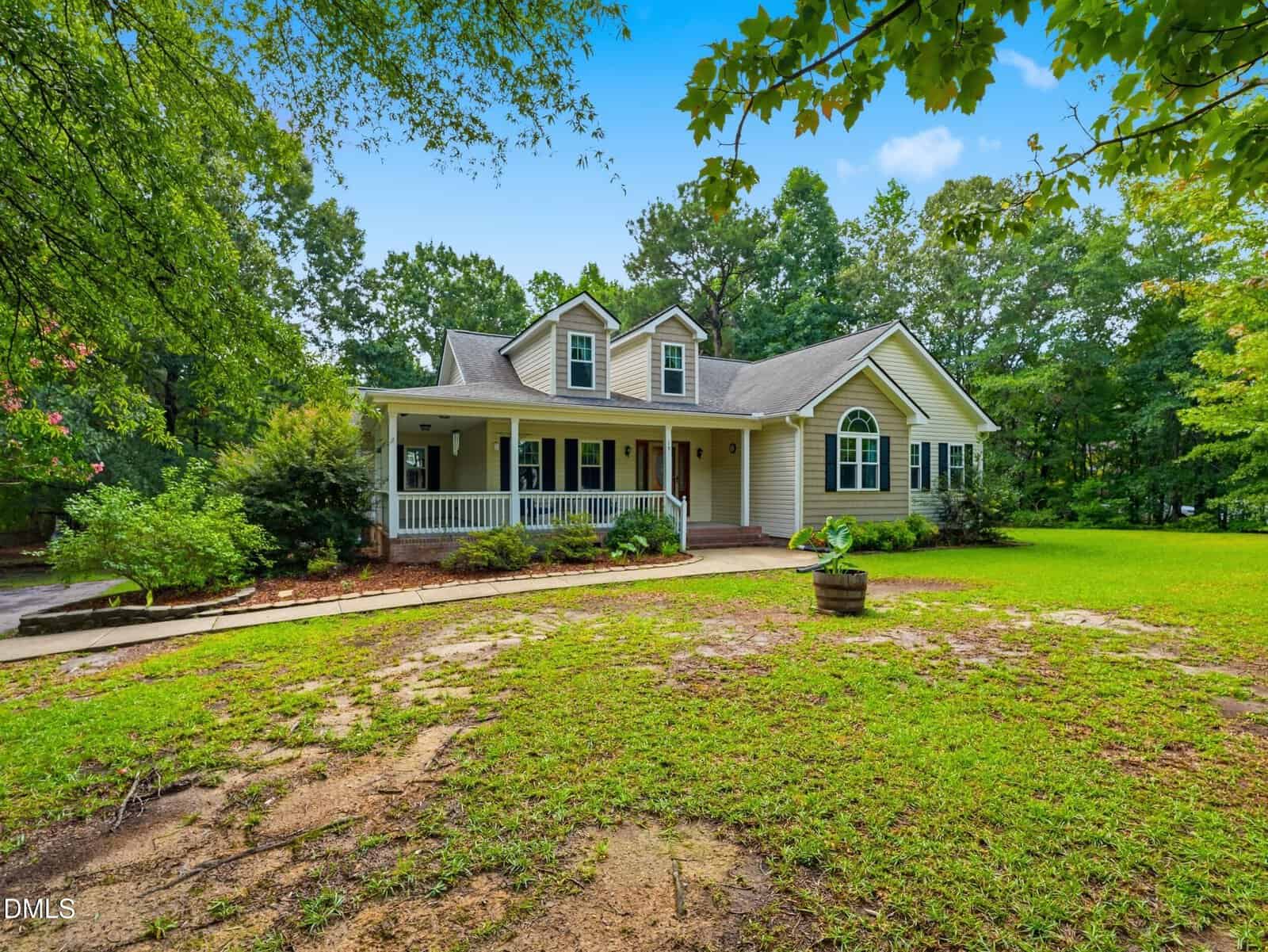 Single-story light gray house with white trim, a covered front porch, dormer windows, and surrounded by green trees and a grassy yard under a bright blue sky.