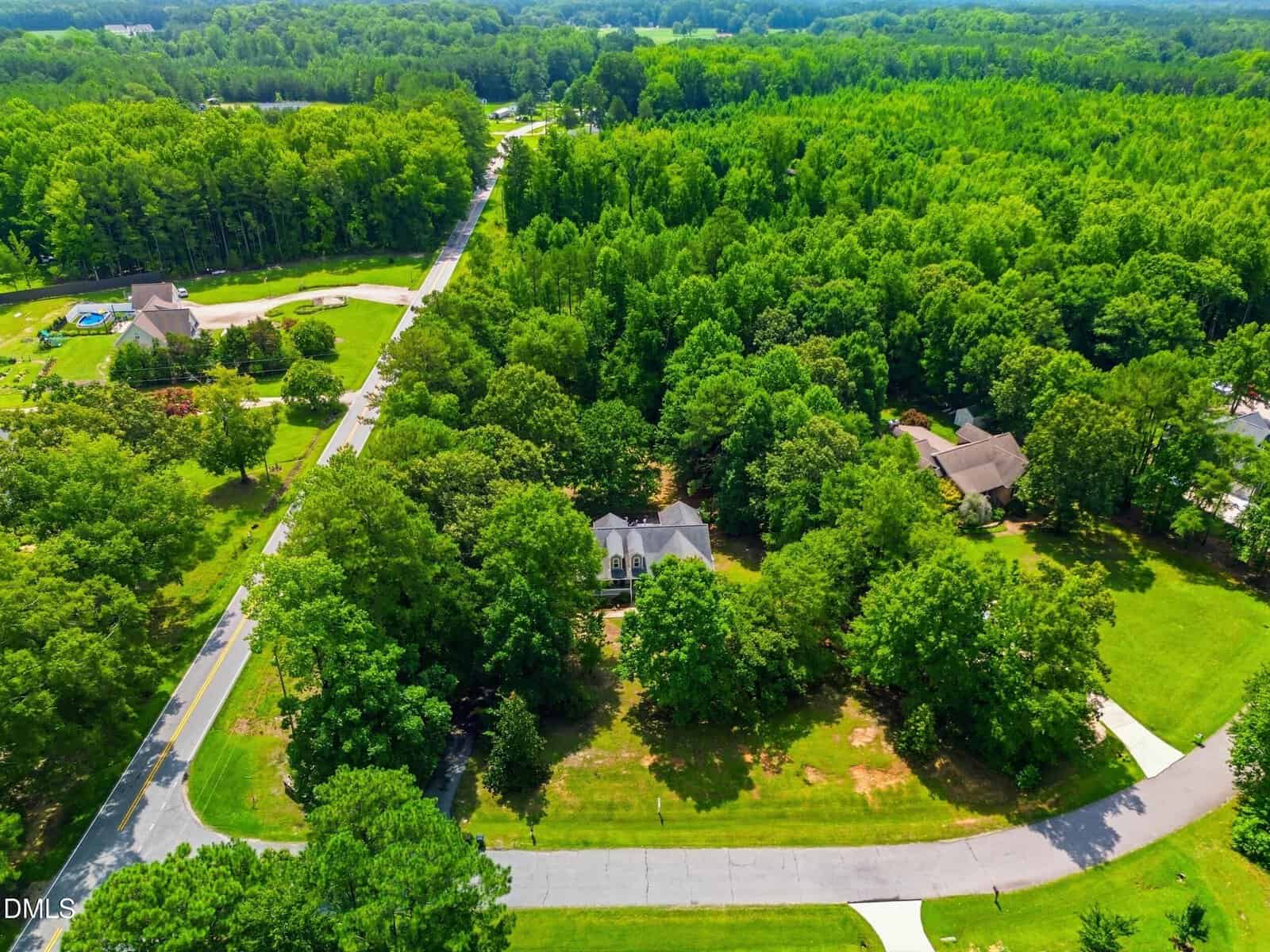 Aerial view of a large house surrounded by trees in a suburban neighborhood with winding roads and nearby homes, all set in a lush, green, wooded area.