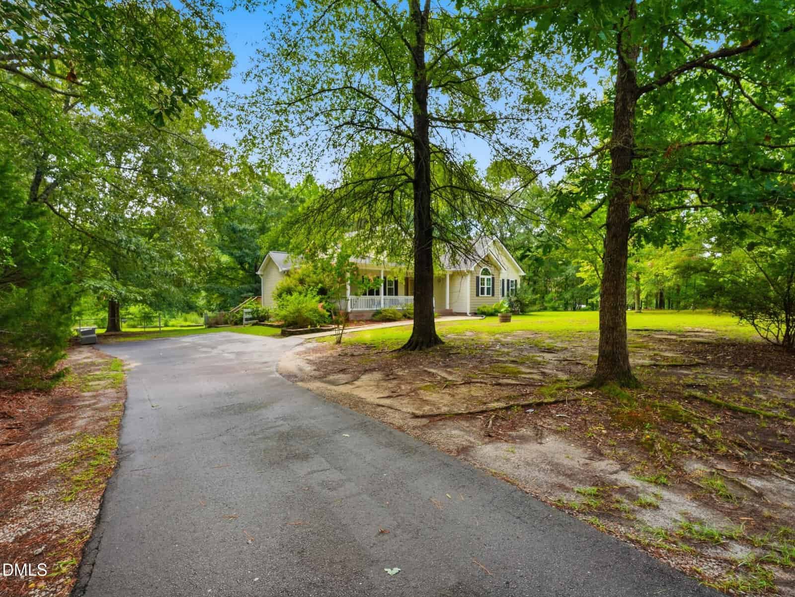 A paved driveway curves toward a light-colored house with a porch, surrounded by trees and green grass on a spacious lot under a bright sky.
