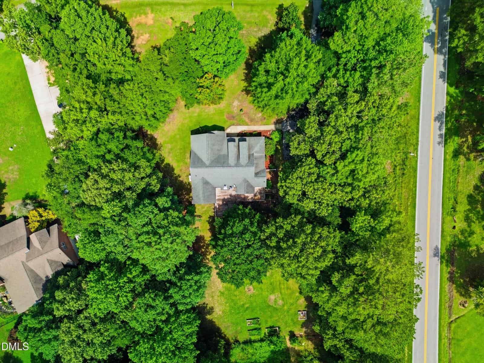 Aerial view of a house surrounded by dense trees, with a road on the right and neighboring houses partially visible. The home has a gray roof and a small yard with green grass and scattered garden beds.