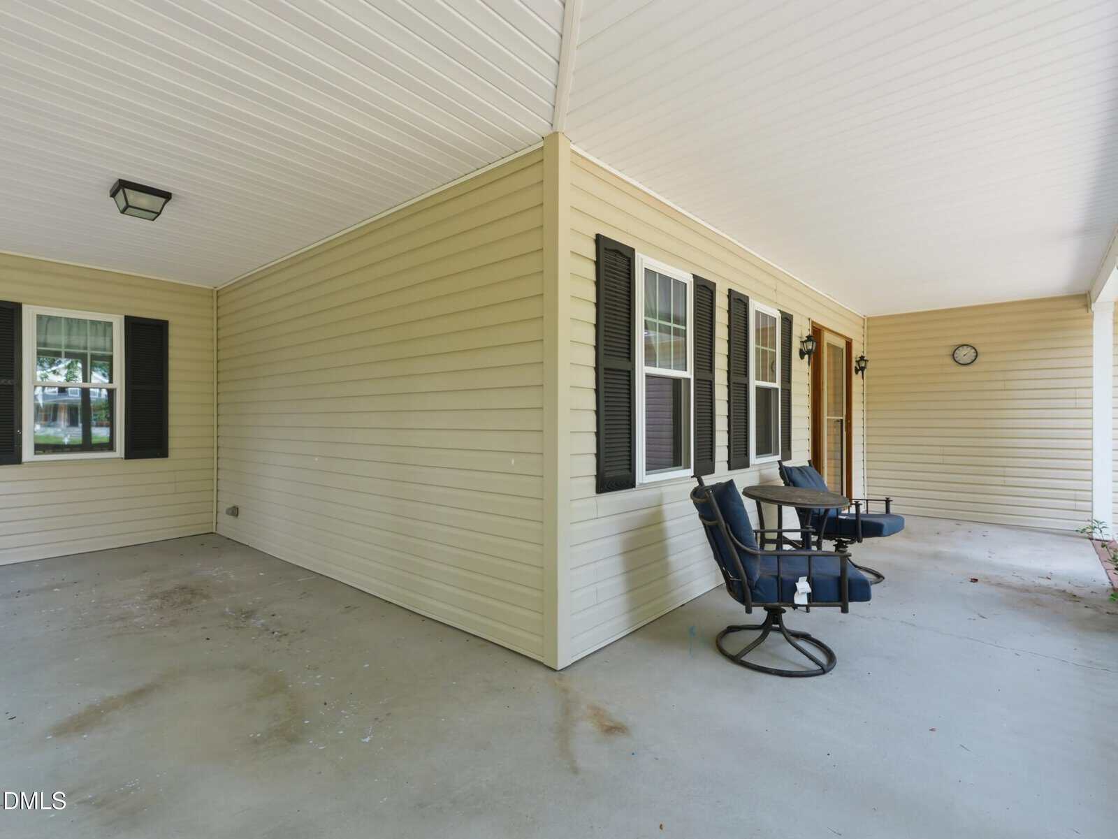 Covered front porch with beige siding, black shutters, two cushioned swivel chairs, a small table, and a wall clock. Large open concrete floor area with windows looking out onto the porch.
