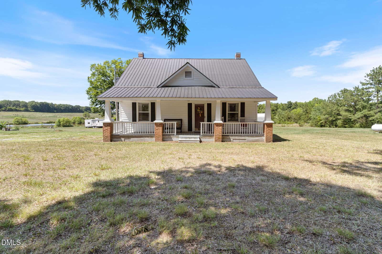 A white farmhouse at 1509 Oak Hill Road, Oxford, with a metal roof and brick porch columns sits in an open grassy yard, surrounded by trees and fields under a blue sky.