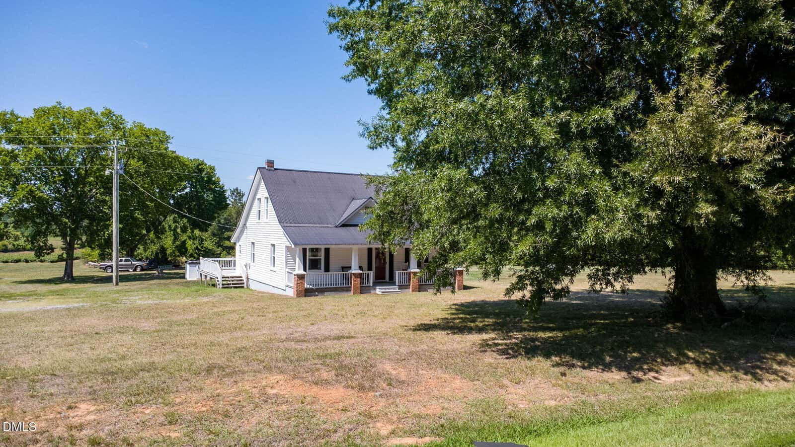 A white house with a porch sits on a large grassy yard, surrounded by trees under a clear blue sky. A car is visible in the background near a dirt driveway.