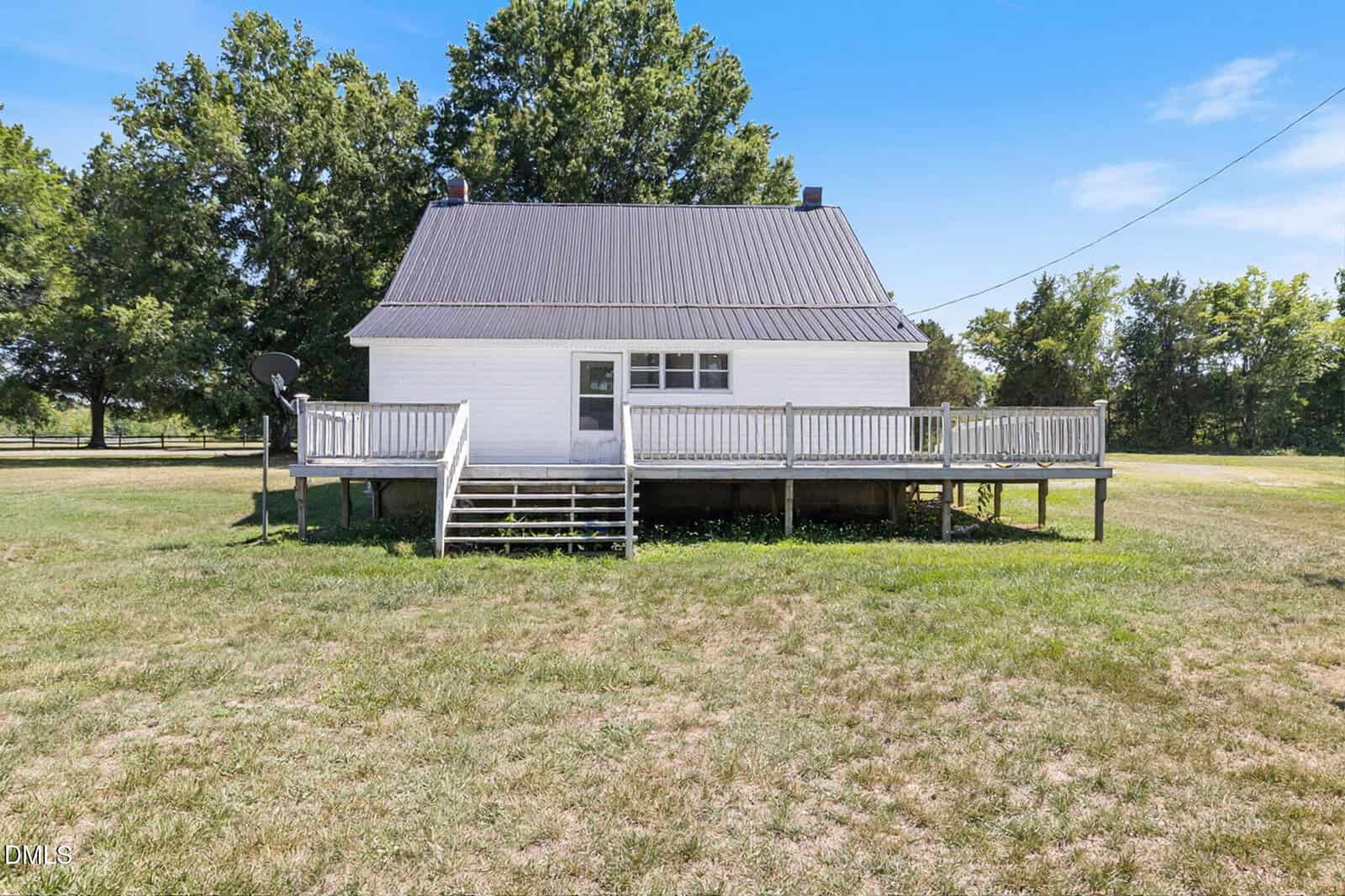 A small white house with a steep metal roof, front porch, and stairs, set on a grassy yard with trees in the background under a clear blue sky.