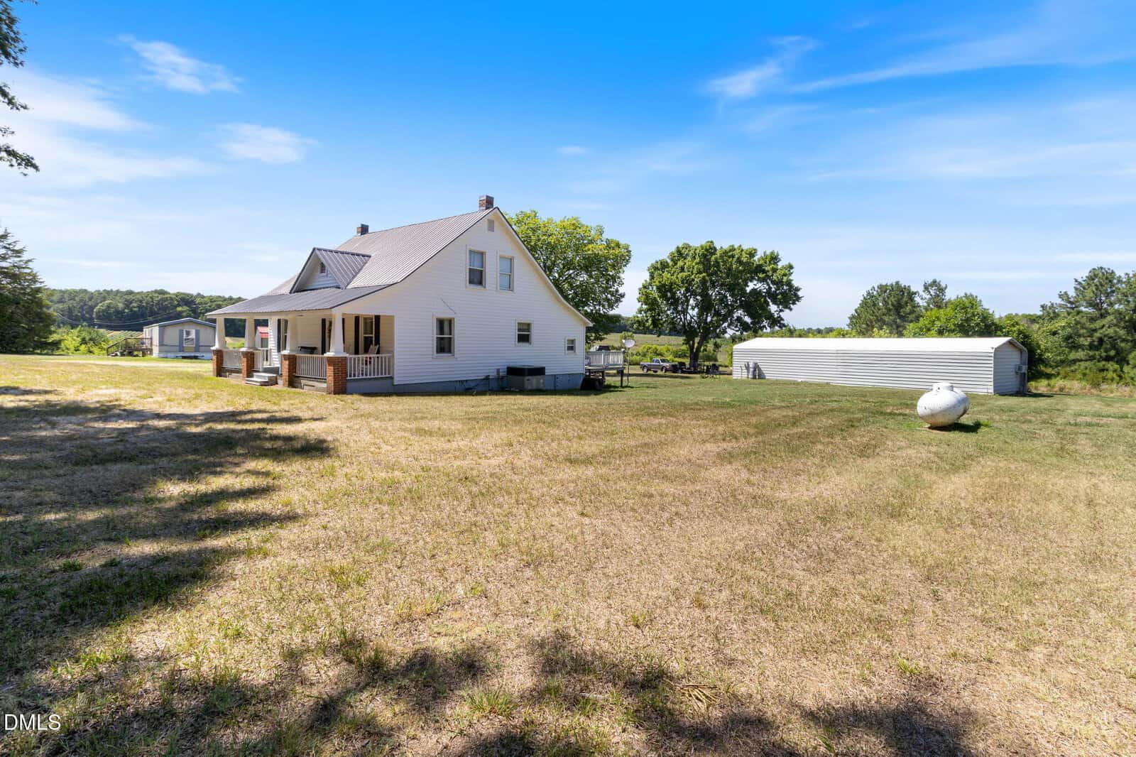 A white house with a covered porch sits on a large grassy lot, with a propane tank and a long white shed in the background under a bright blue sky.