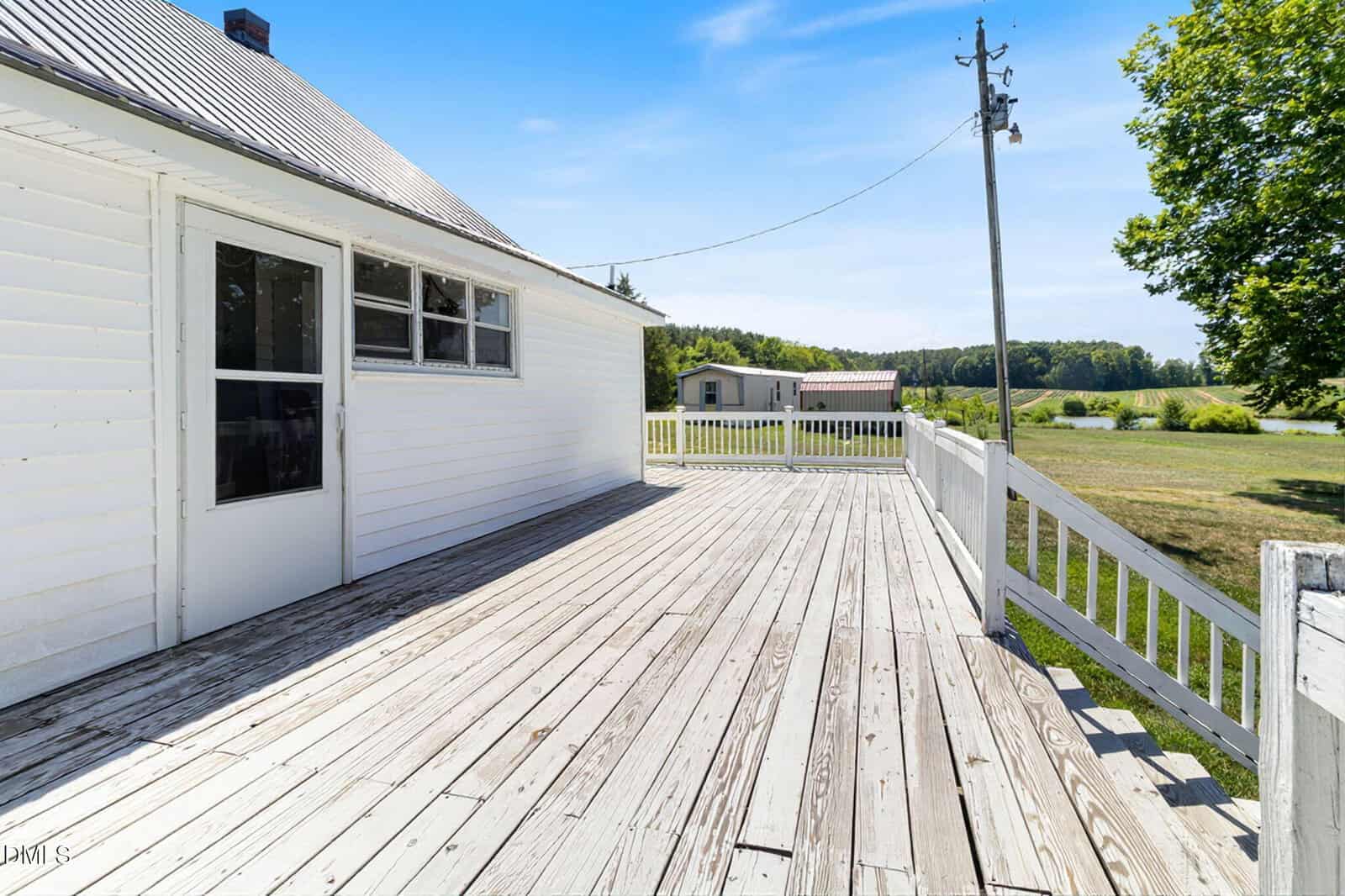 A spacious wooden deck attached to a white house overlooks a grassy yard, trees, and a distant building under a clear blue sky.