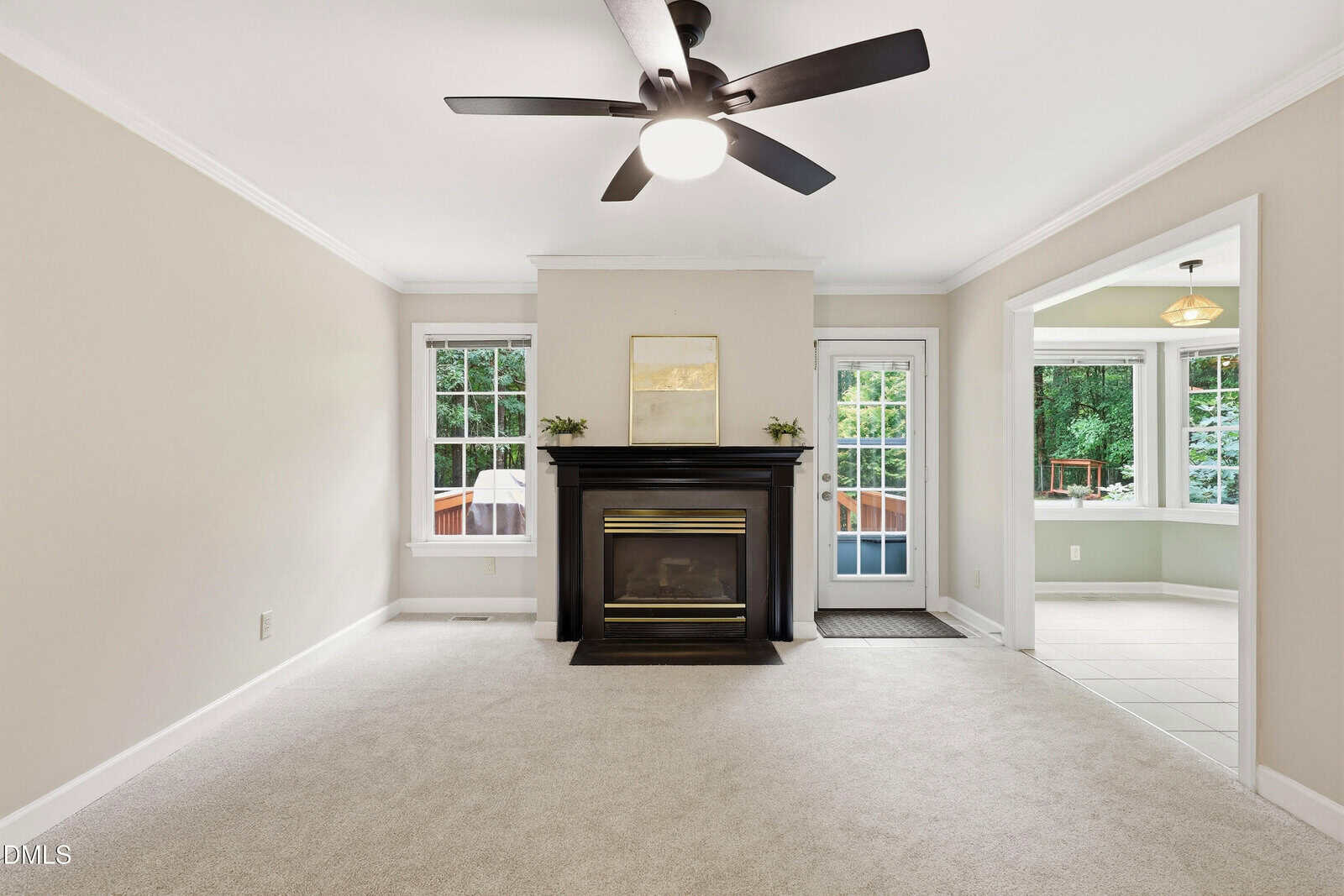 Bright living room with beige walls, carpeted floor, black fireplace, ceiling fan, two windows, and a glass door leading to a sunroom with tile flooring and large windows facing greenery.