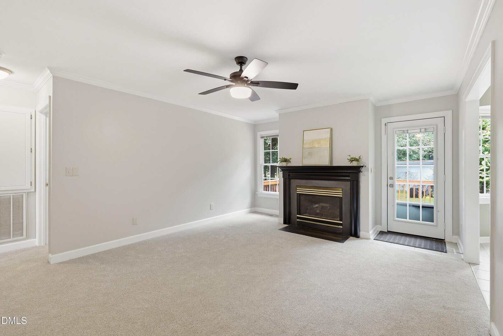 Bright living room with light walls and carpet, a ceiling fan, black fireplace with a painting above, two windows, and a glass door leading outside to a green backyard.