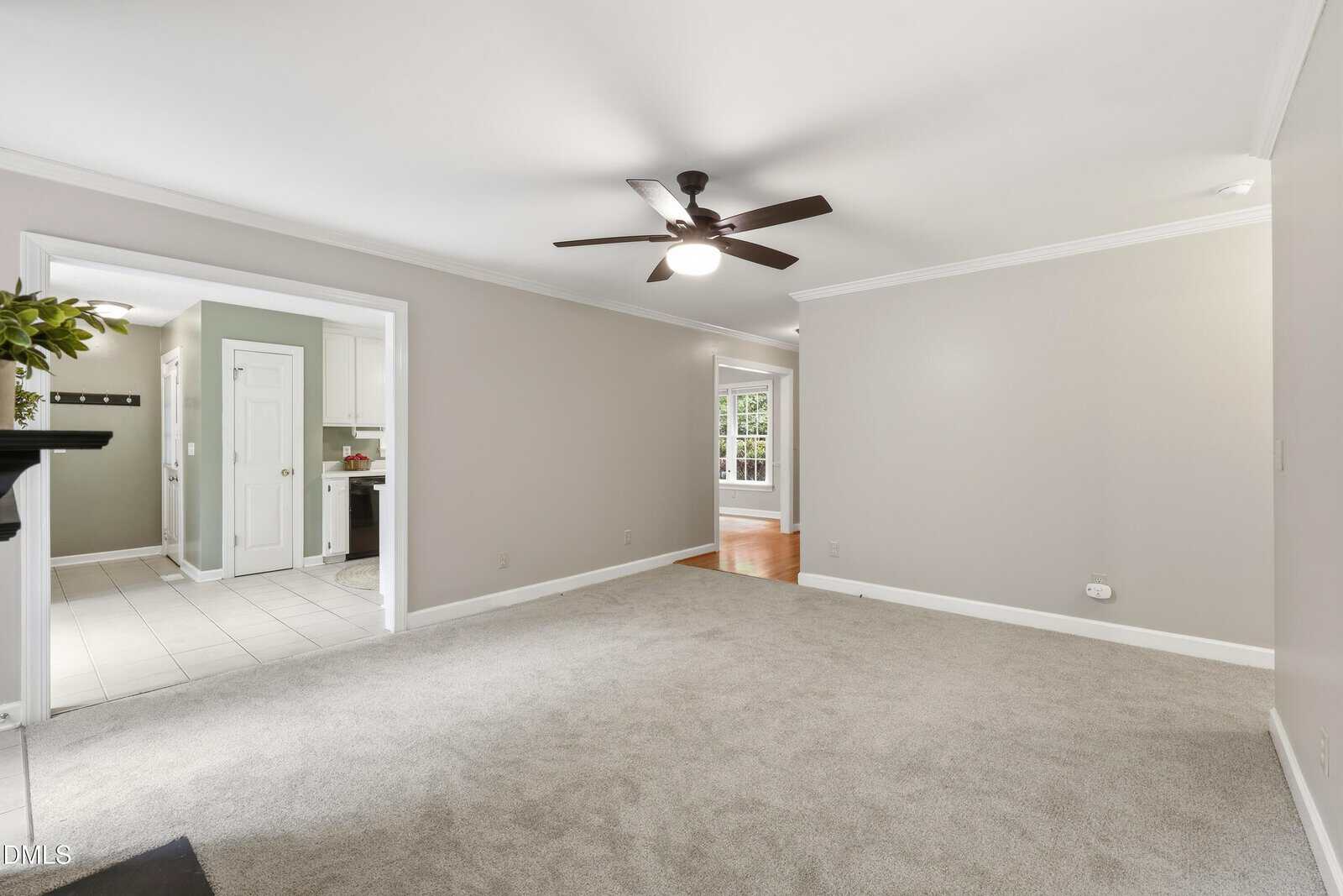 A bright, carpeted living room with light gray walls, ceiling fan, crown molding, and open doorways to a tiled kitchen and a hardwood dining area. Large windows let in natural light.