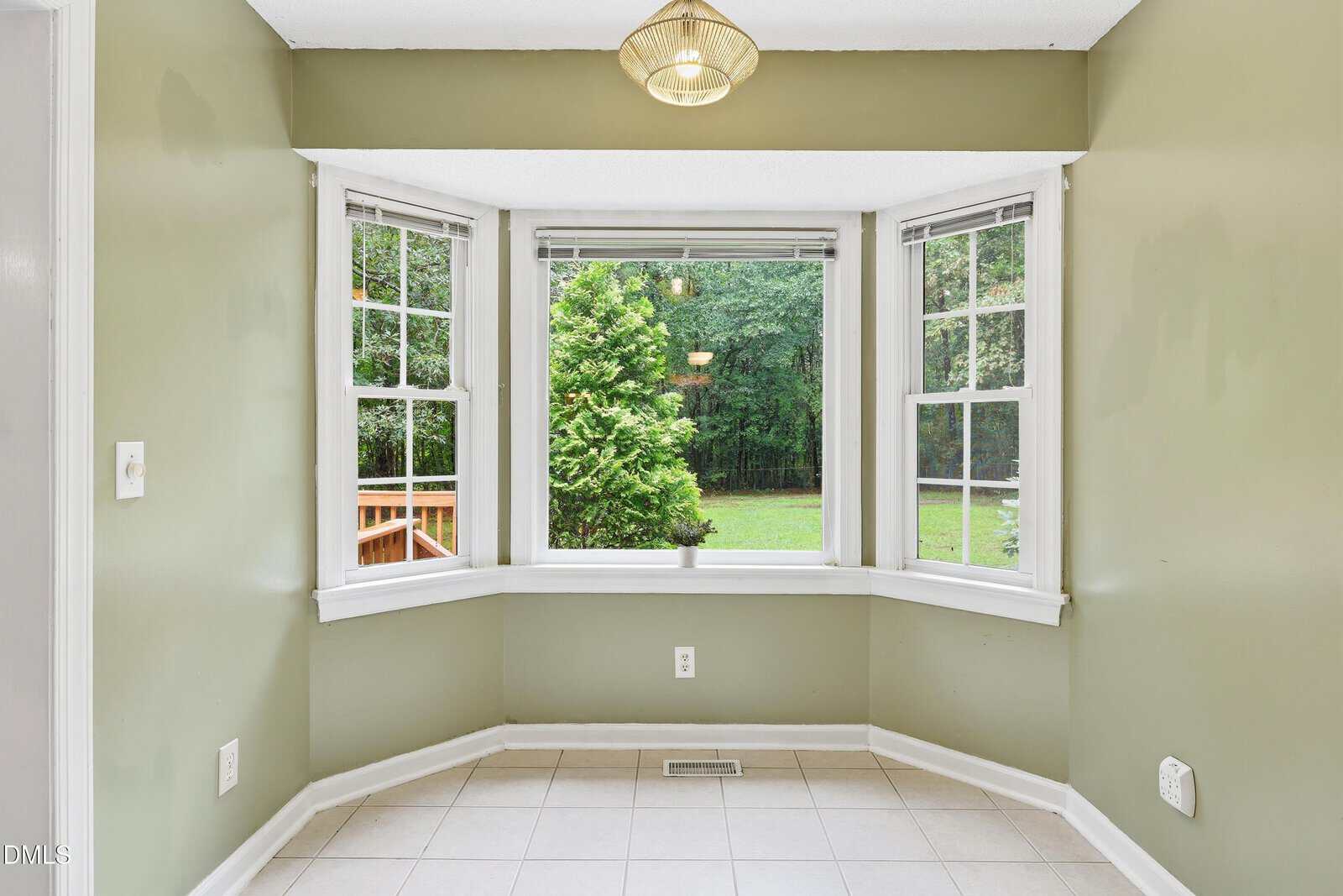 A bay window with three sections overlooks a green backyard. The room has sage-green walls, white trim, a tiled floor, and a ceiling light fixture. A wooden deck is visible outside to the left.