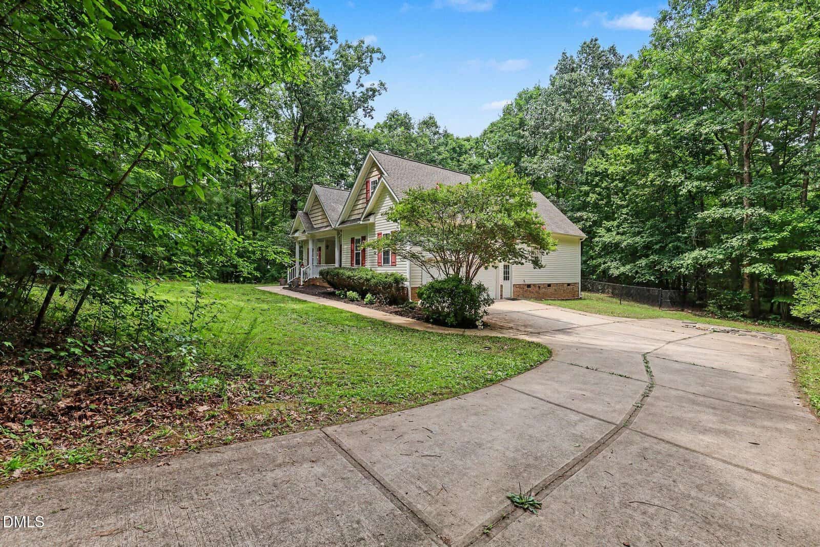 A single-story house with white siding, a front porch, and gabled roof sits beside a curved driveway, surrounded by tall trees and greenery on a clear day.