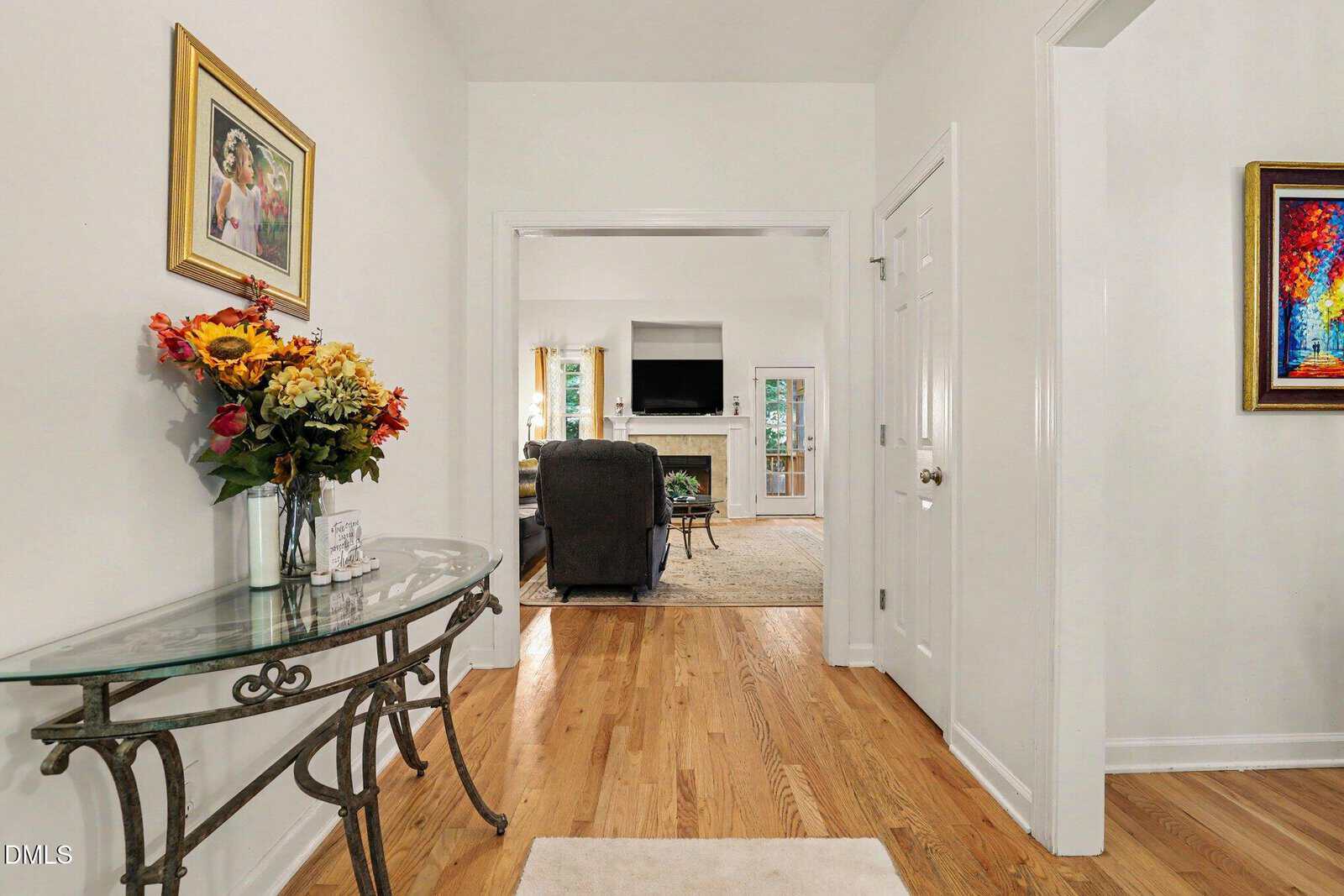 A hallway with hardwood floors, a glass-top table with a flower vase, and framed art leads to a living room with armchairs, a fireplace, and a TV. Natural light comes through windows in the background.
