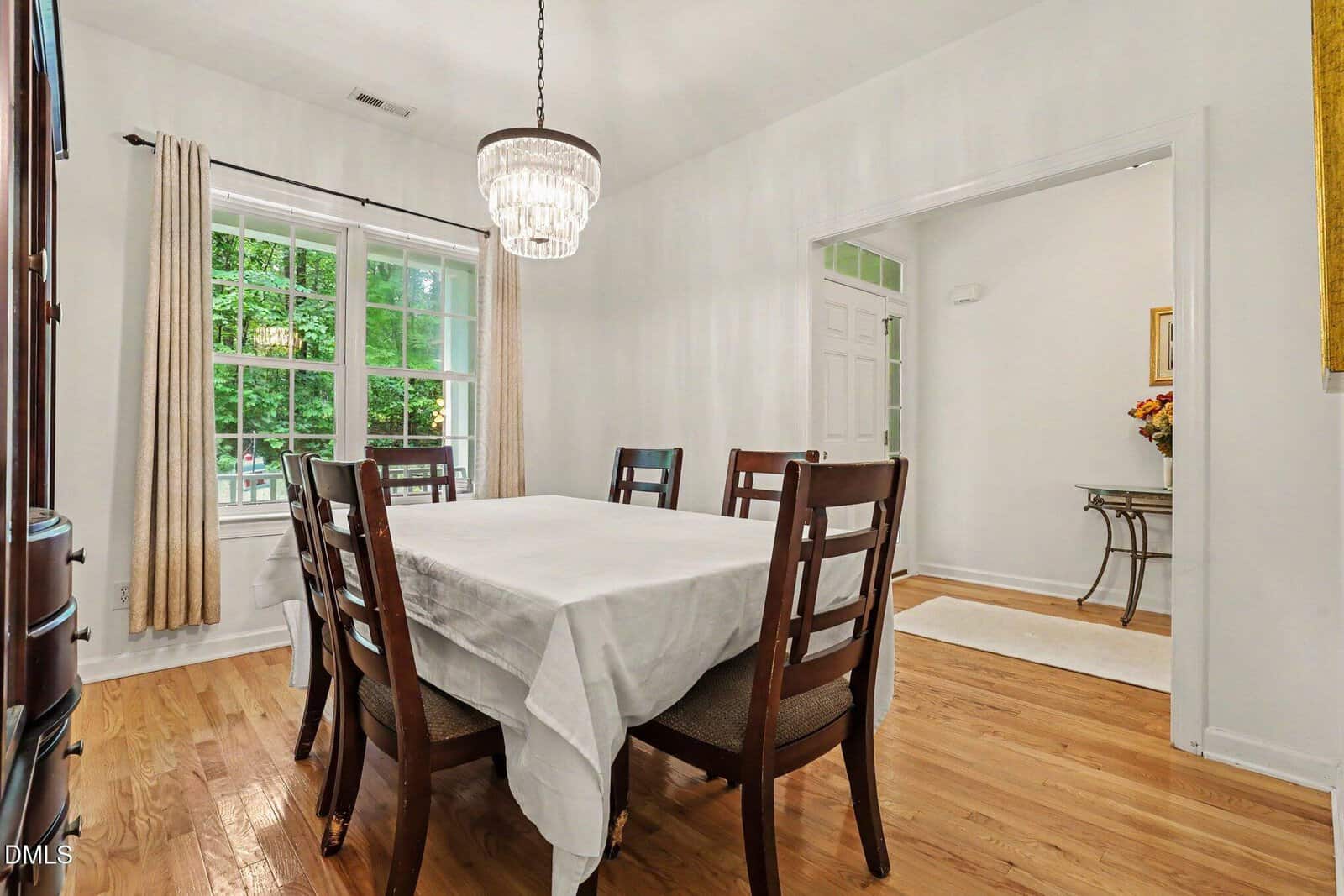 A dining room with a wooden table covered by a white tablecloth, six chairs, a chandelier, hardwood floors, and large windows with beige curtains. An open doorway leads to a bright entryway with a table and flowers.