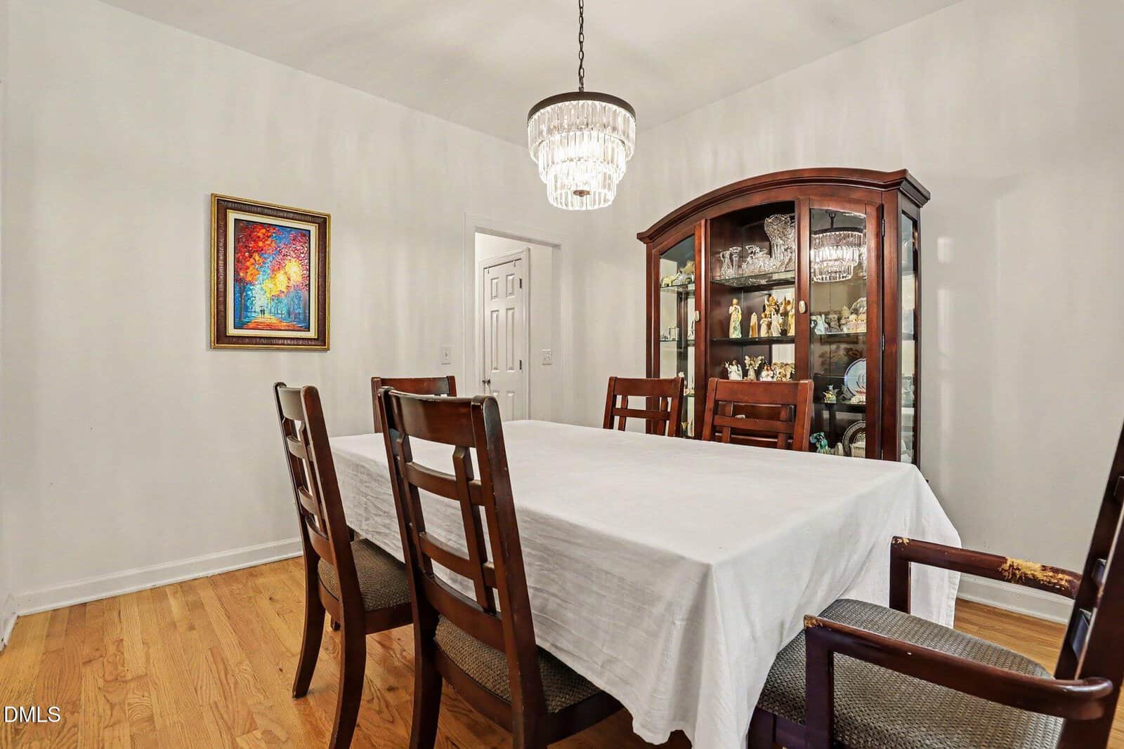 A dining room with a wooden table covered by a white cloth, six chairs, a glass-fronted china cabinet, a crystal chandelier, and a colorful painting on a white wall.