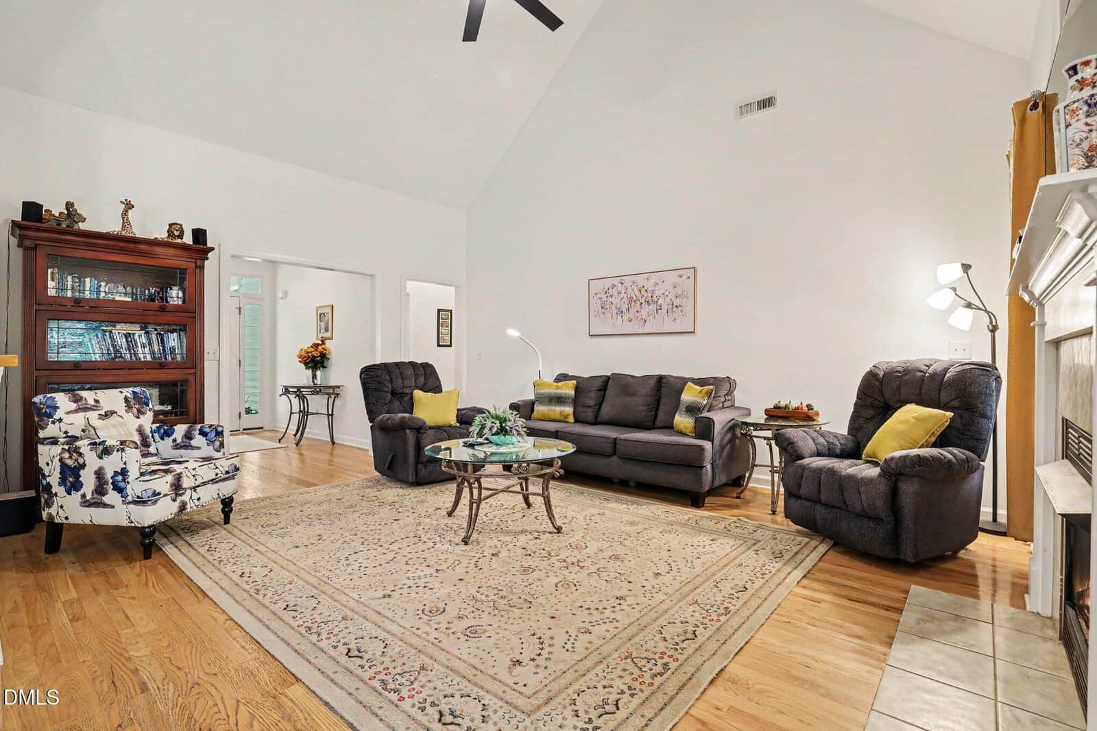 Spacious living room with a patterned rug, gray sofas, a floral armchair, glass coffee table, wooden bookshelf, and modern floor lamp. Light walls and wood flooring create a bright, welcoming atmosphere.