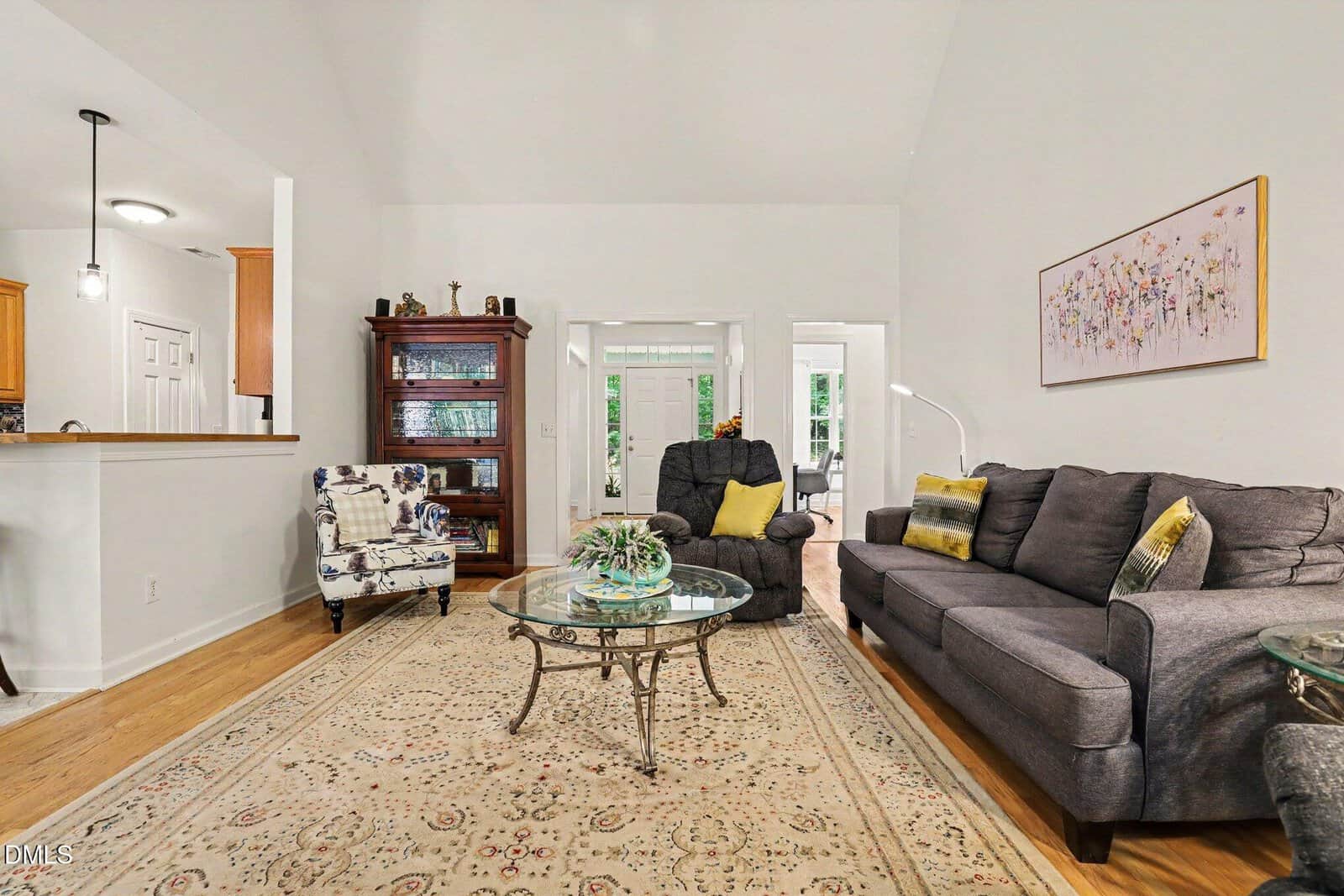 A cozy living room with a gray sofa, floral armchair, glass coffee tables, an ornate rug, wooden floor, a china cabinet, and a floral painting on the wall. Bright light from doors at the back illuminates the space.