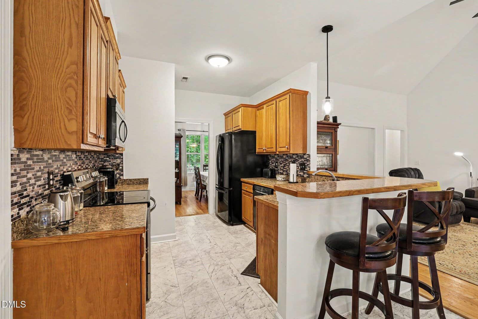 Modern kitchen with wooden cabinets, black appliances, tiled backsplash, marble floor, and a breakfast bar with two dark wooden chairs. An open doorway leads to a dining area with large windows in the background.