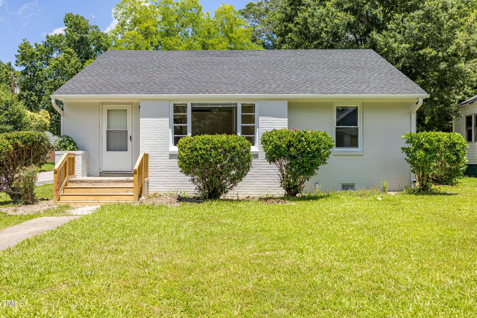 Charming small white house with a dark roof at 605 E Montgomery Street, Henderson, featuring a central front window, wooden steps leading to the door on the left, and neatly trimmed bushes and green lawn in front. Trees and blue sky provide a picturesque background.
