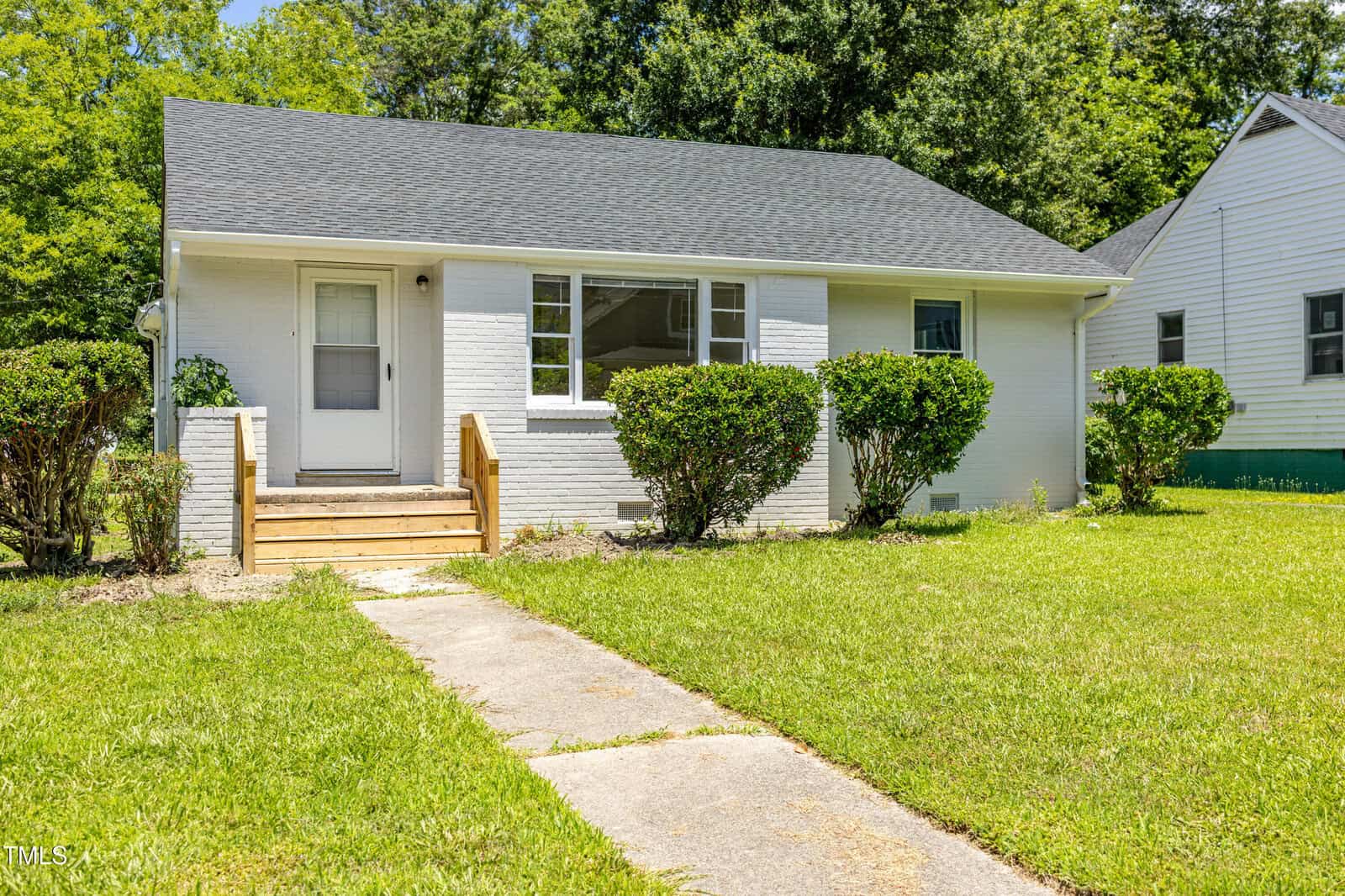 A small white brick house with a dark roof, front steps, and shrubs by the entrance. The yard is grassy with a concrete walkway leading to the door. Trees and neighboring homes are visible in the background.