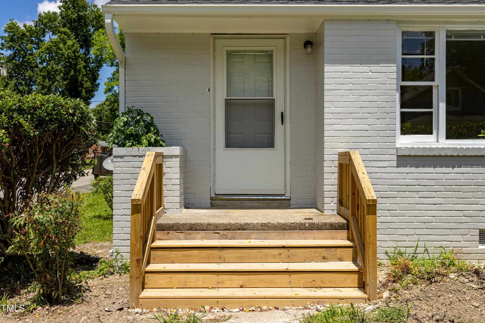 A small concrete porch with three wooden steps and railings leads to a white door on a light gray brick house. There are green plants on both sides of the steps and a window to the right of the door.
