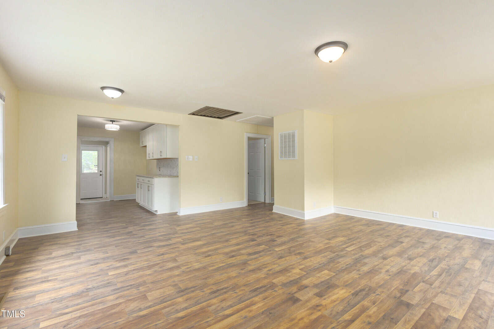 Spacious, empty room with wood flooring, cream-colored walls, and ceiling lights. An open doorway leads to a kitchen area with white cabinets and a back door. Multiple windows provide natural light.