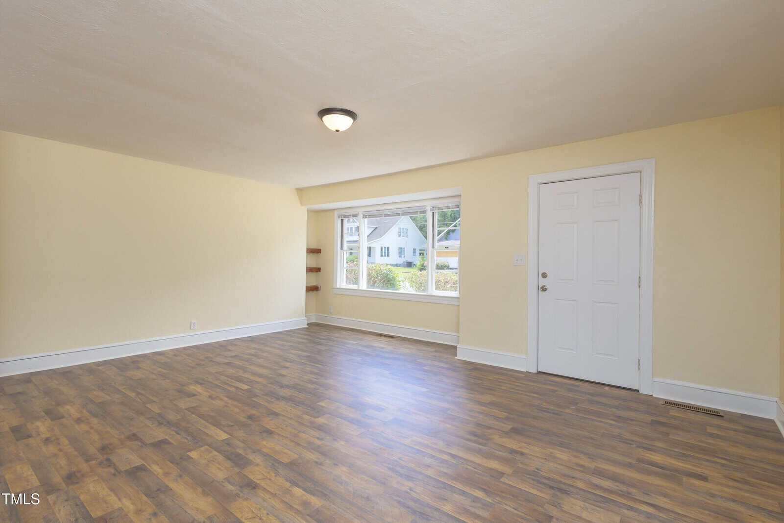 Empty room with light yellow walls, wood flooring, a ceiling light, a large window letting in natural light, and a white door. A few houses and greenery are visible outside the window.