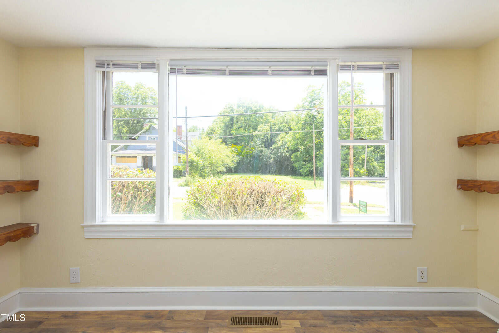 Large three-panel window with white trim in a beige room, featuring wooden shelves on both sides and a view of green shrubs and trees outside. Sunlight fills the space, highlighting the wood floor and open, airy feel.