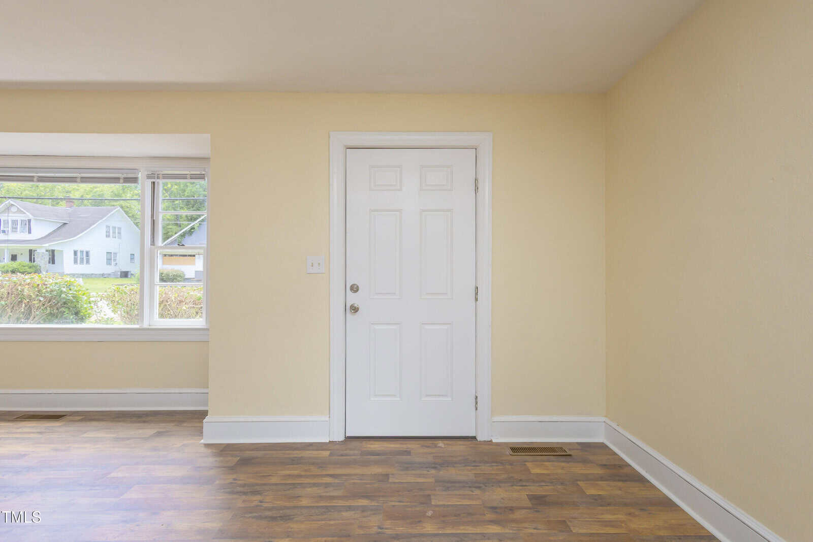 A white front door with a round handle is centered in a cream-colored room with wood flooring. To the left, a large window lets in natural light and shows houses and greenery outside.