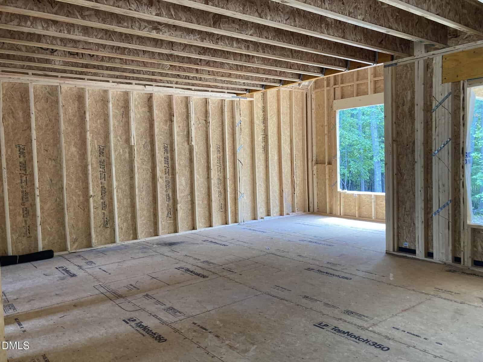 Interior of a house under construction showing exposed wooden framing, unfinished walls, and a large window opening with trees visible outside. Natural light fills the space from the window.