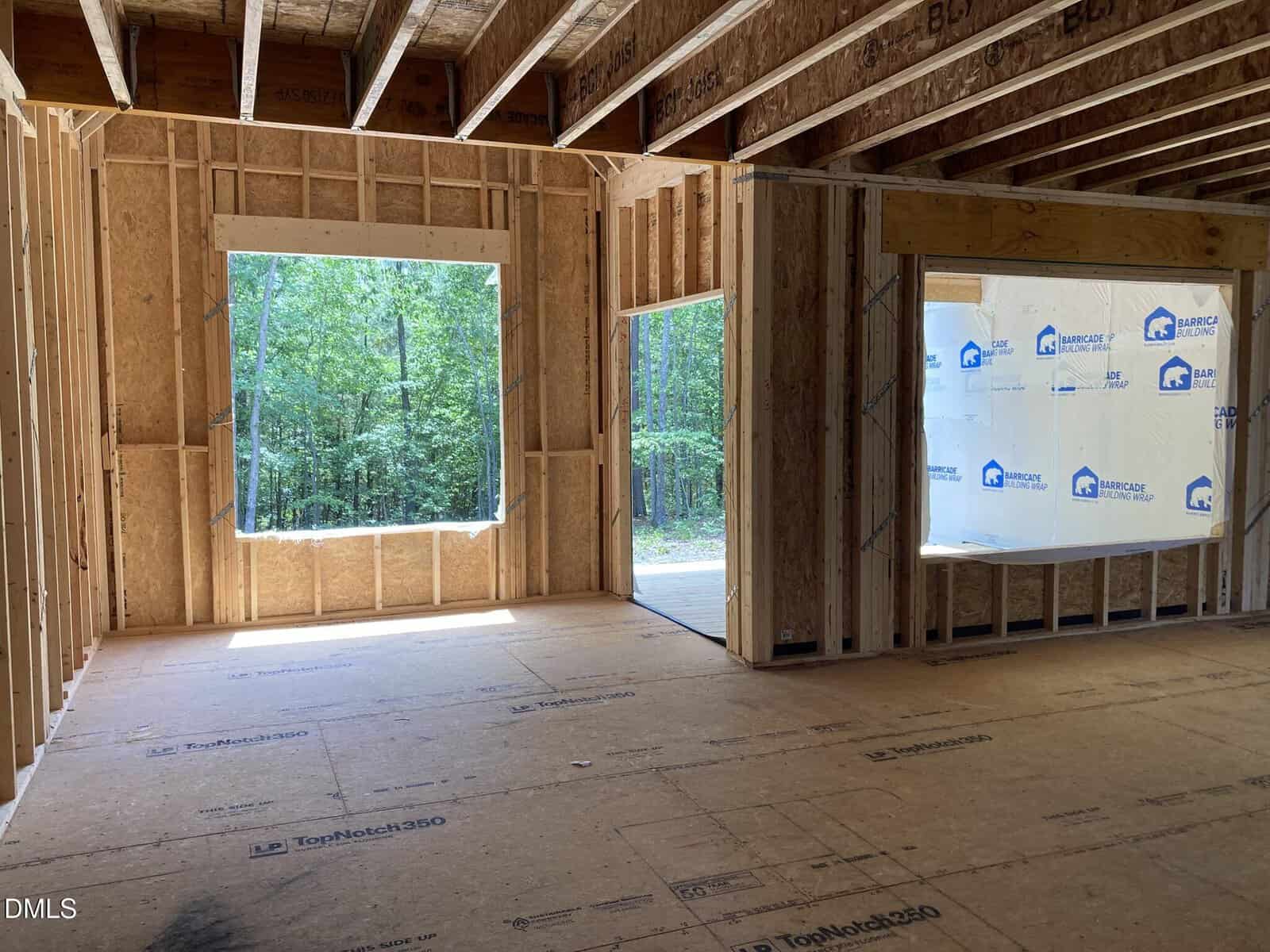 Interior of a house under construction with exposed wooden framing, large window openings, and sunlight streaming in. Forested trees are visible outside the windows.
